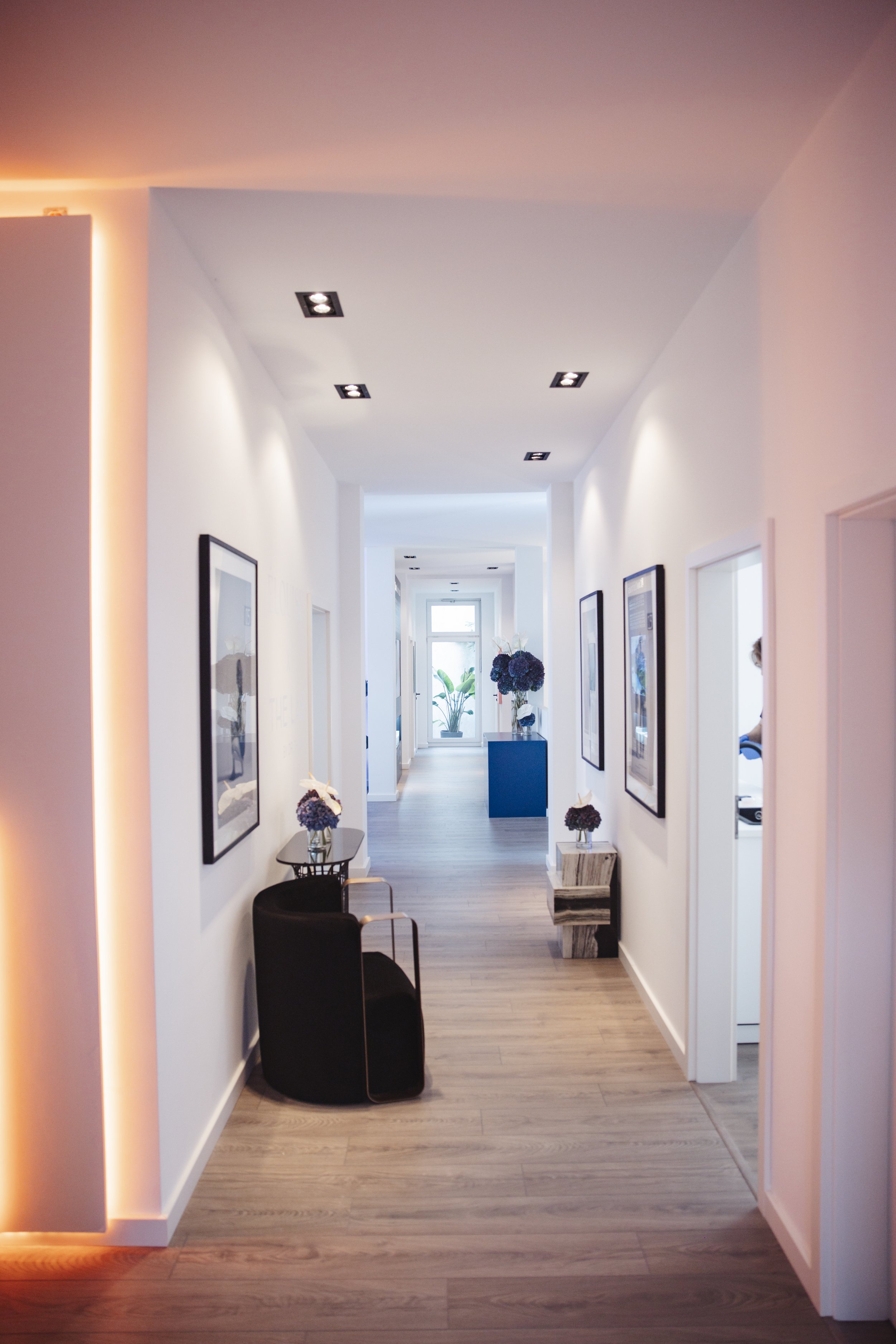 Ein heller, moderner Flur in einer Praxis für Plastische & Ästhetische Chirurgie mit Kosmetikinstitut mit weißen Wänden, Holzfußboden, Wanddekorationen und einem Fenster im Hintergrund, mit Blumenarrangements auf Tischen und einem blauen Sideboard.
