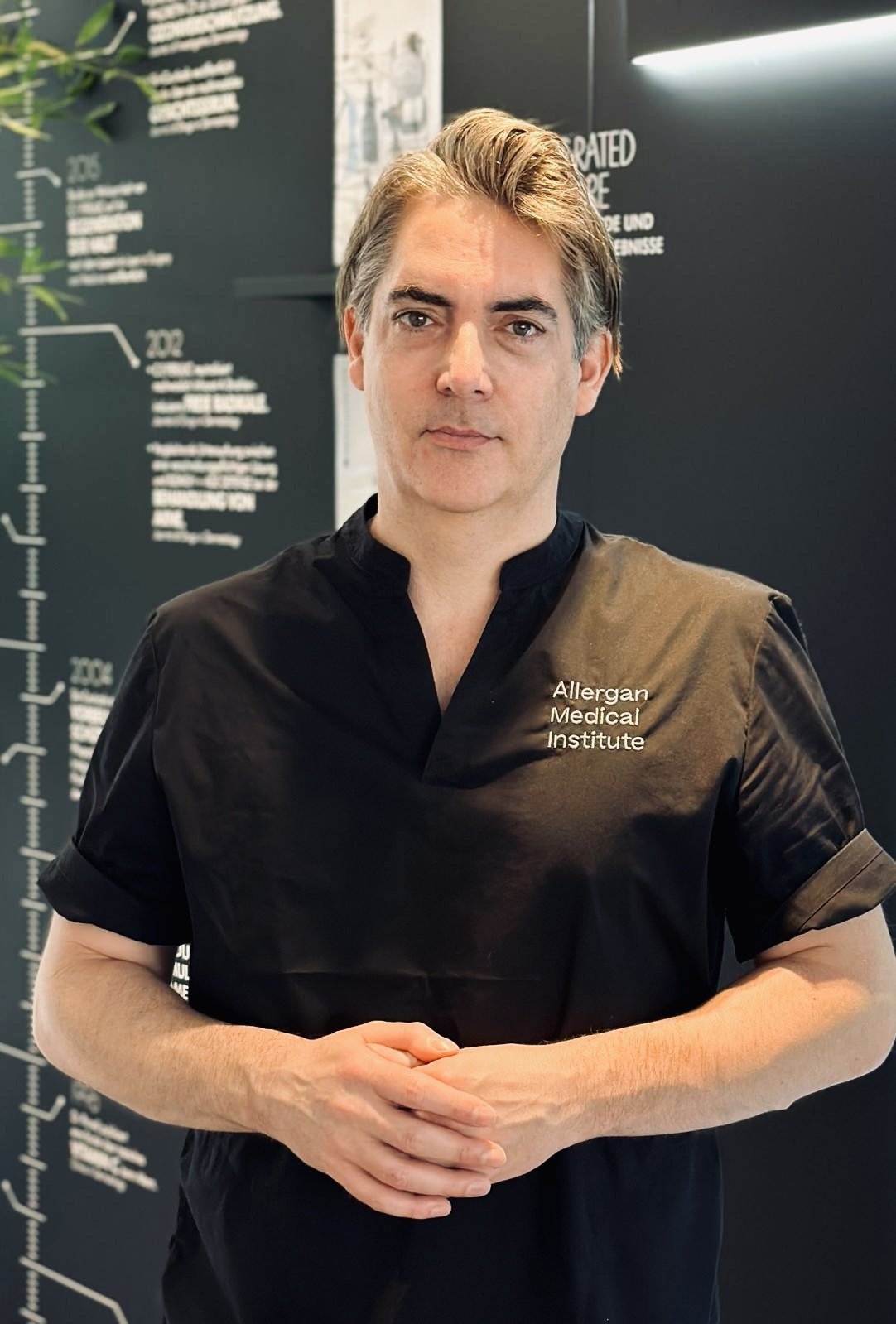 Ein Mann trägt ein schwarzes medizinisches Kleid mit der Aufschrift 'Allergan Medical Institute' und steht vor einer schwarzen Wand mit medizinischen Informationen und Diagrammen in einer Klinik oder einem medizinischen Institut.