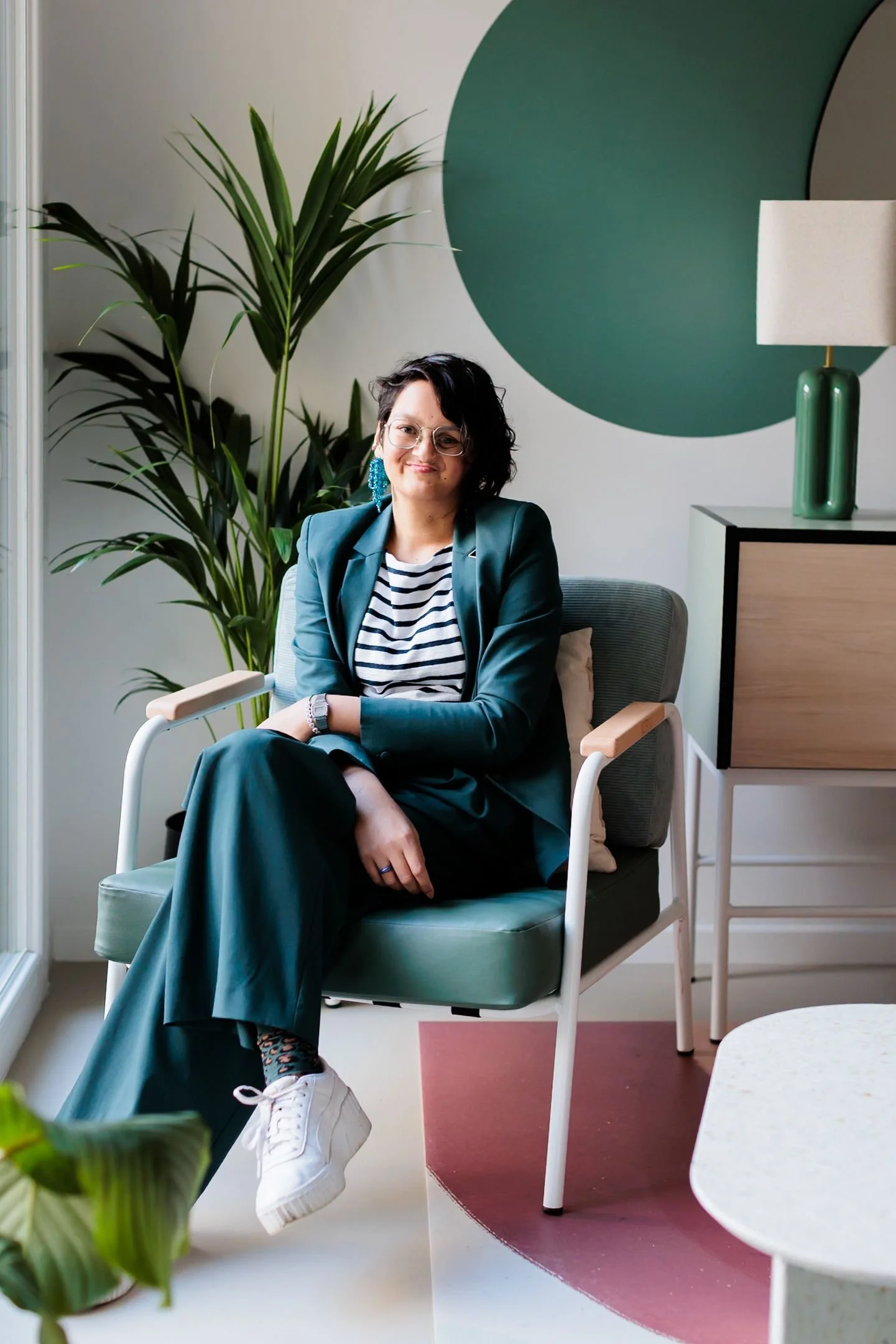 Une femme assise dans un fauteuil, portant une veste verte et un t-shirt rayé, dans un intérieur moderne avec une grande plante verte et un tableau abstrait vert sur le mur.