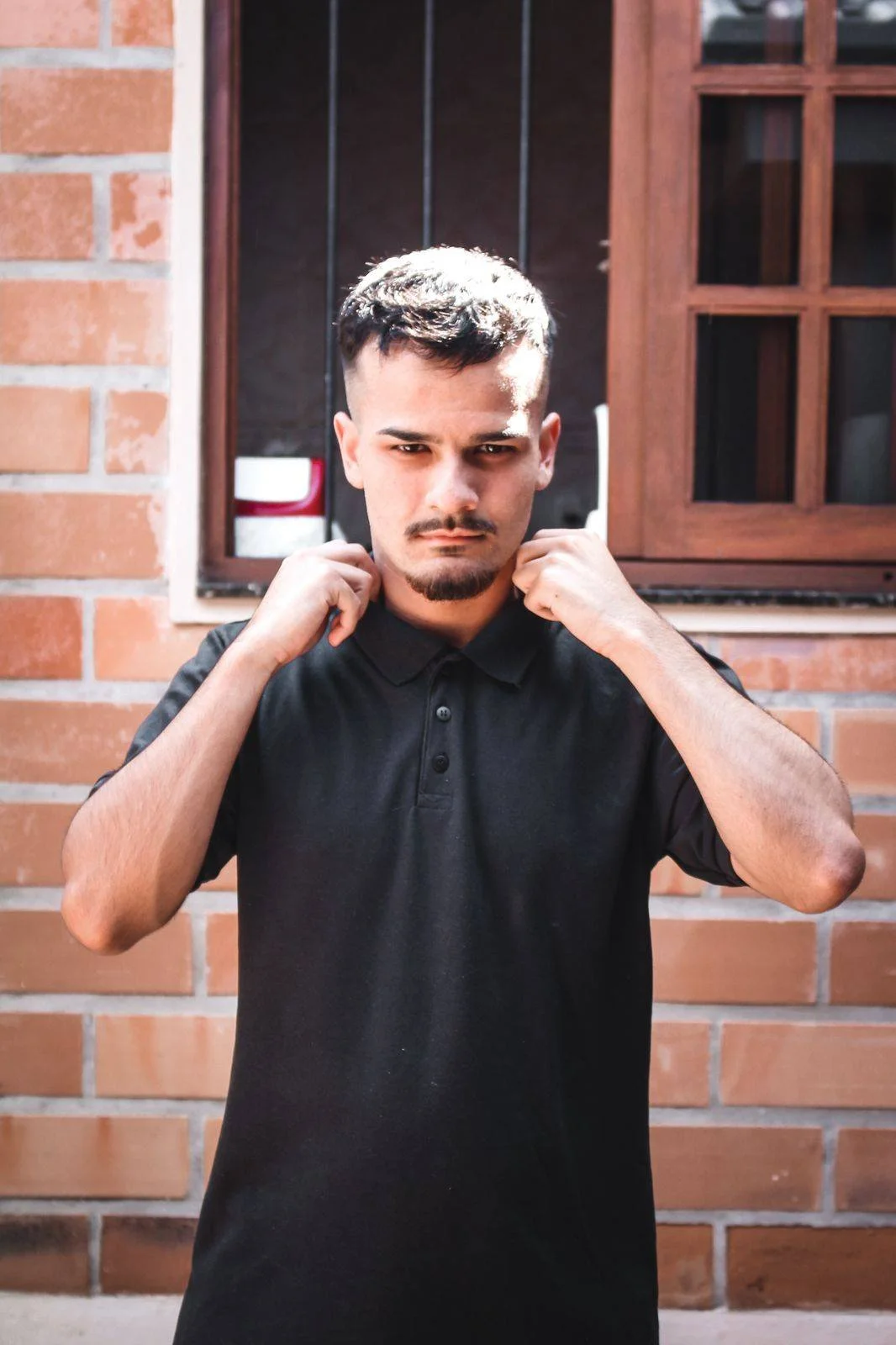 Young man adjusting his collar in front of a brick wall and window.
