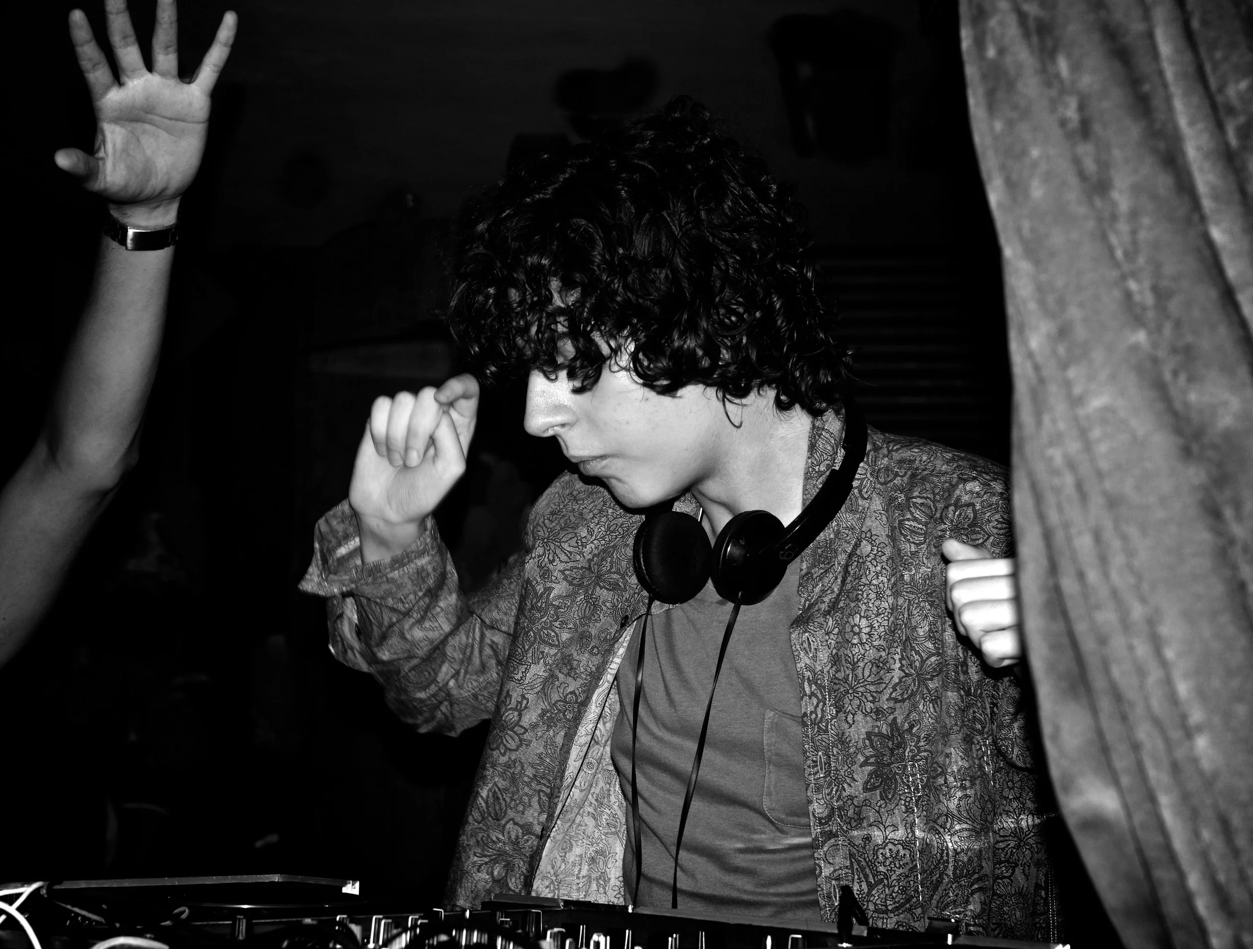 A person with curly hair wearing headphones and a patterned shirt, appears to be DJing or mixing music at a club or event, with a hand raised nearby in the foreground.