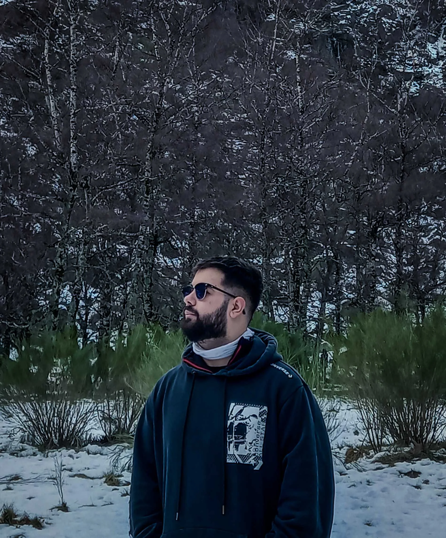 A man with dark hair and beard wearing sunglasses, a face mask under his chin, and a dark hoodie, standing in a snowy outdoor area with trees and bushes in the background.