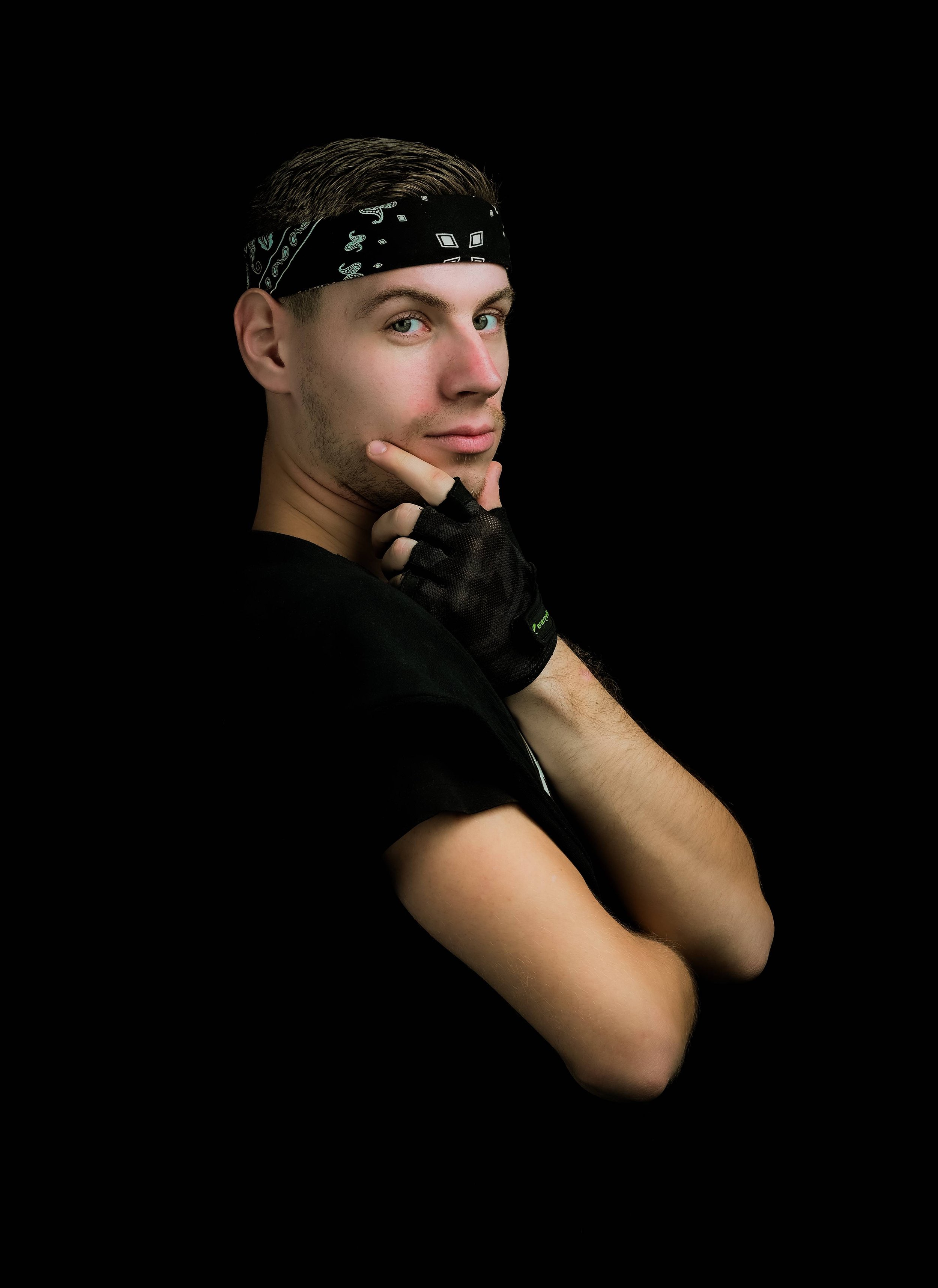 A young man with light skin, blue eyes, and short brown hair wearing a black bandana with white patterns, a black shirt, and black gloves, posing against a black background with his hand on his chin.