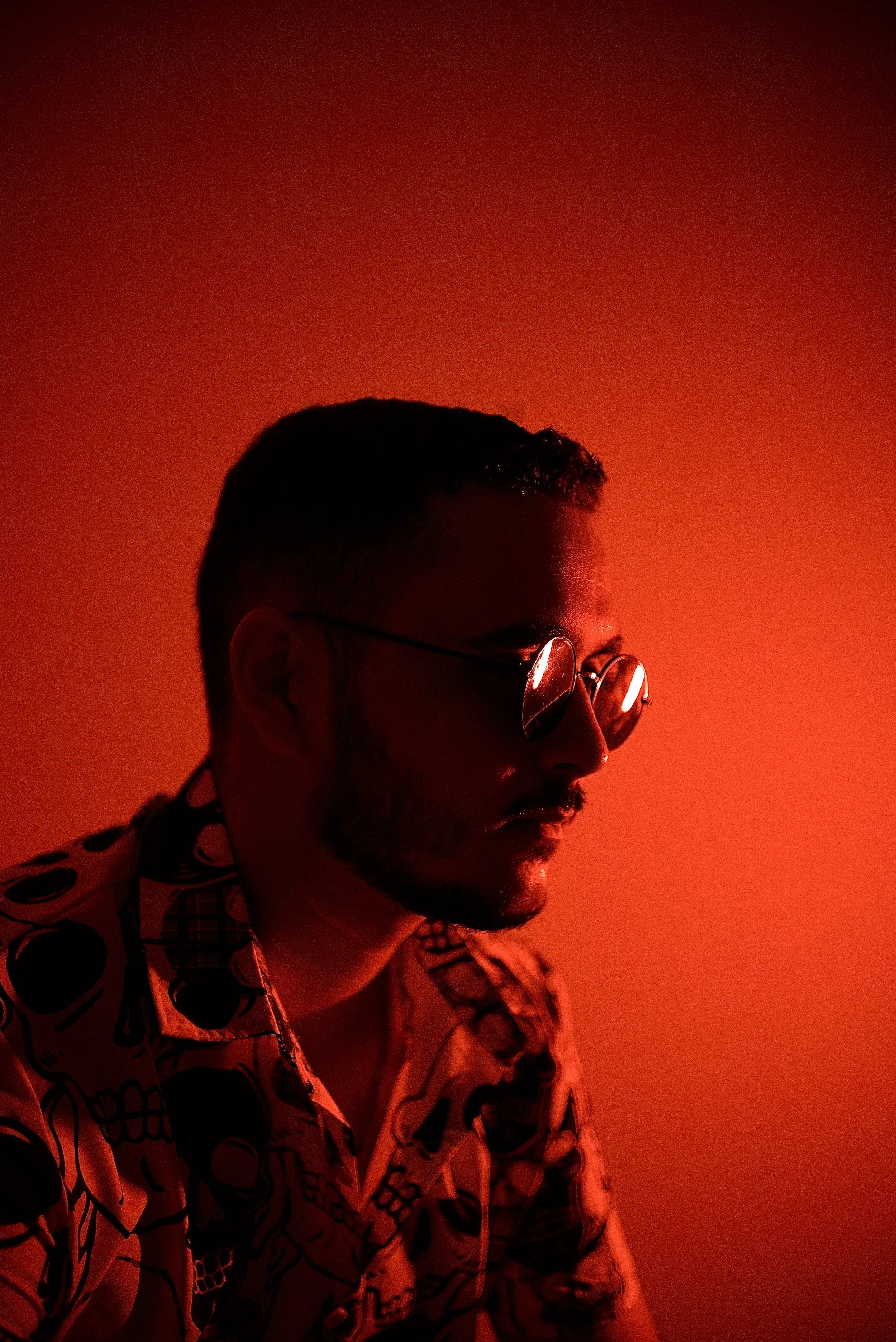 Side profile of a man wearing sunglasses and a patterned shirt, illuminated by red background light.