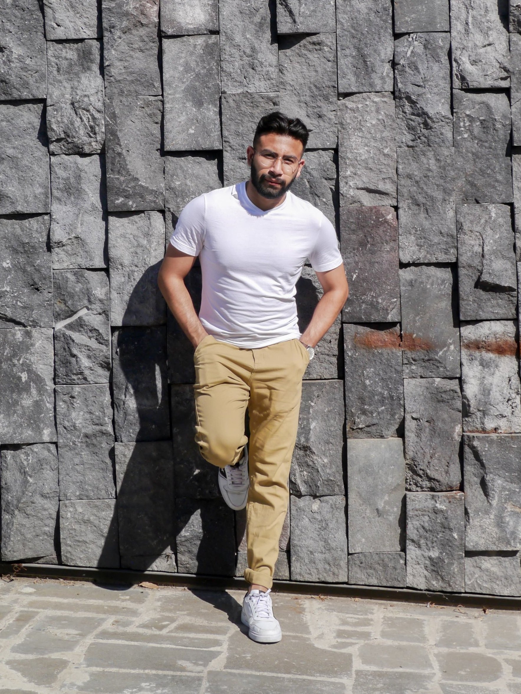 A man with dark hair and beard, wearing glasses, a white t-shirt, beige pants, and white sneakers, leaning against a stone wall with his hands in his pockets, looking at the camera.