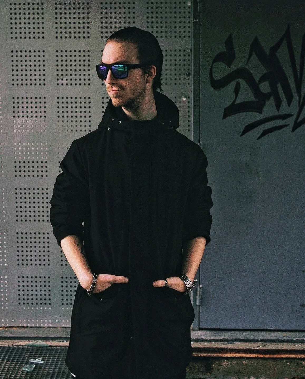 A young man with a braid, sunglasses, and a black hoodie standing against a perforated metal wall with graffiti.