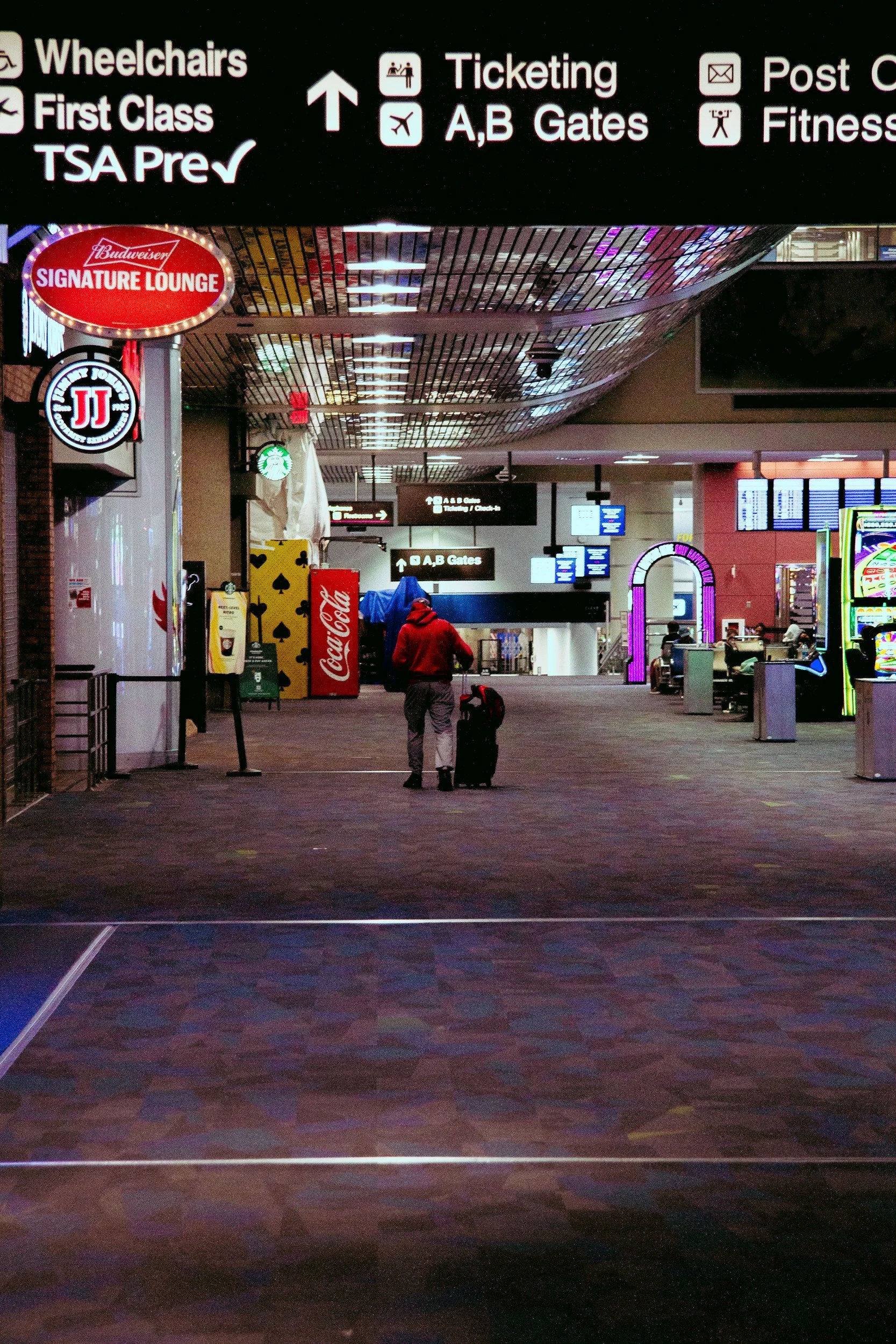 Empty airport terminal with a person pulling luggage, illuminated signs showing gates A and B, and various advertisements and signage.