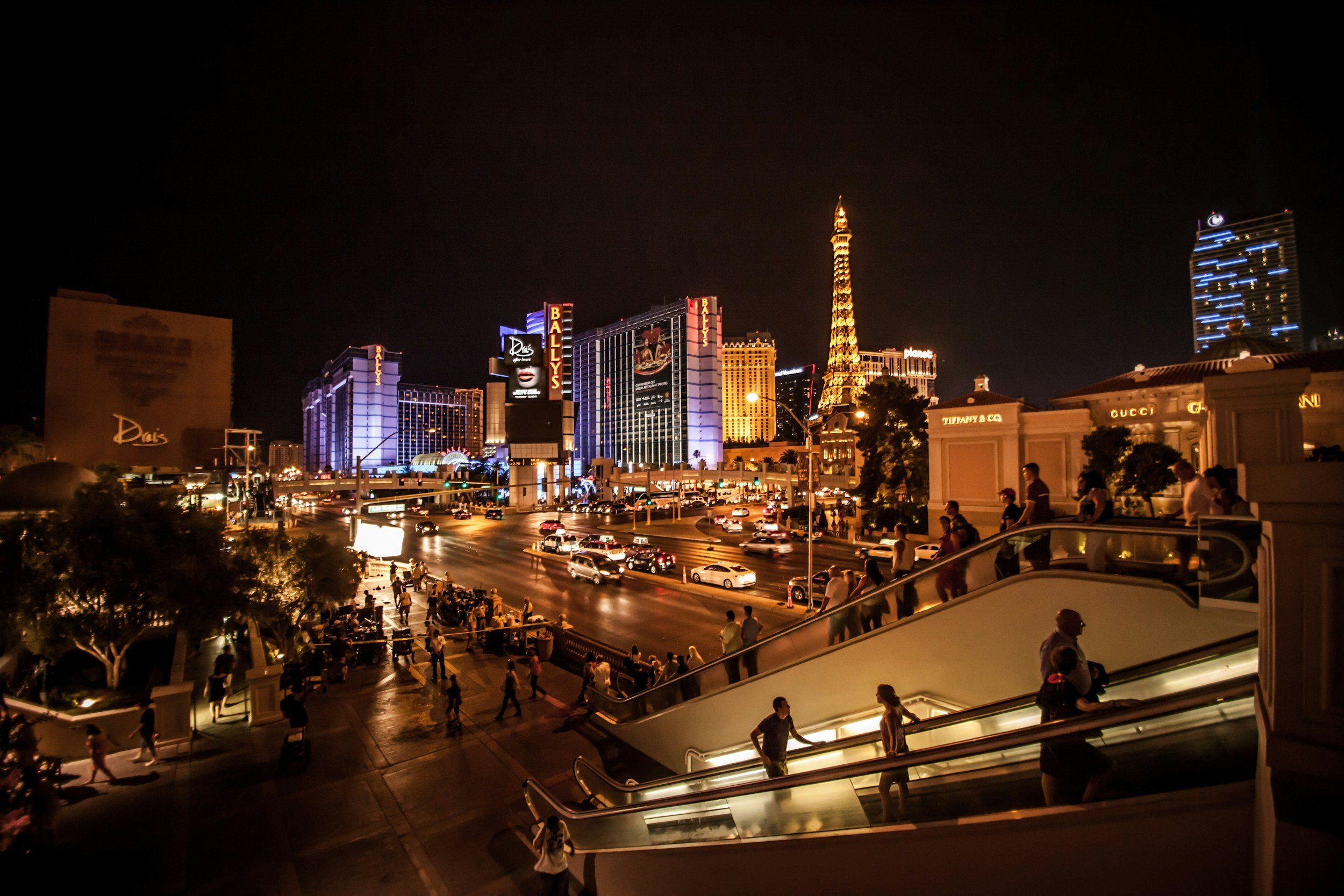 Nighttime view of Las Vegas strip with illuminated hotels and landmarks, including the Eiffel Tower replica, crowds walking, and a busy street with cars.