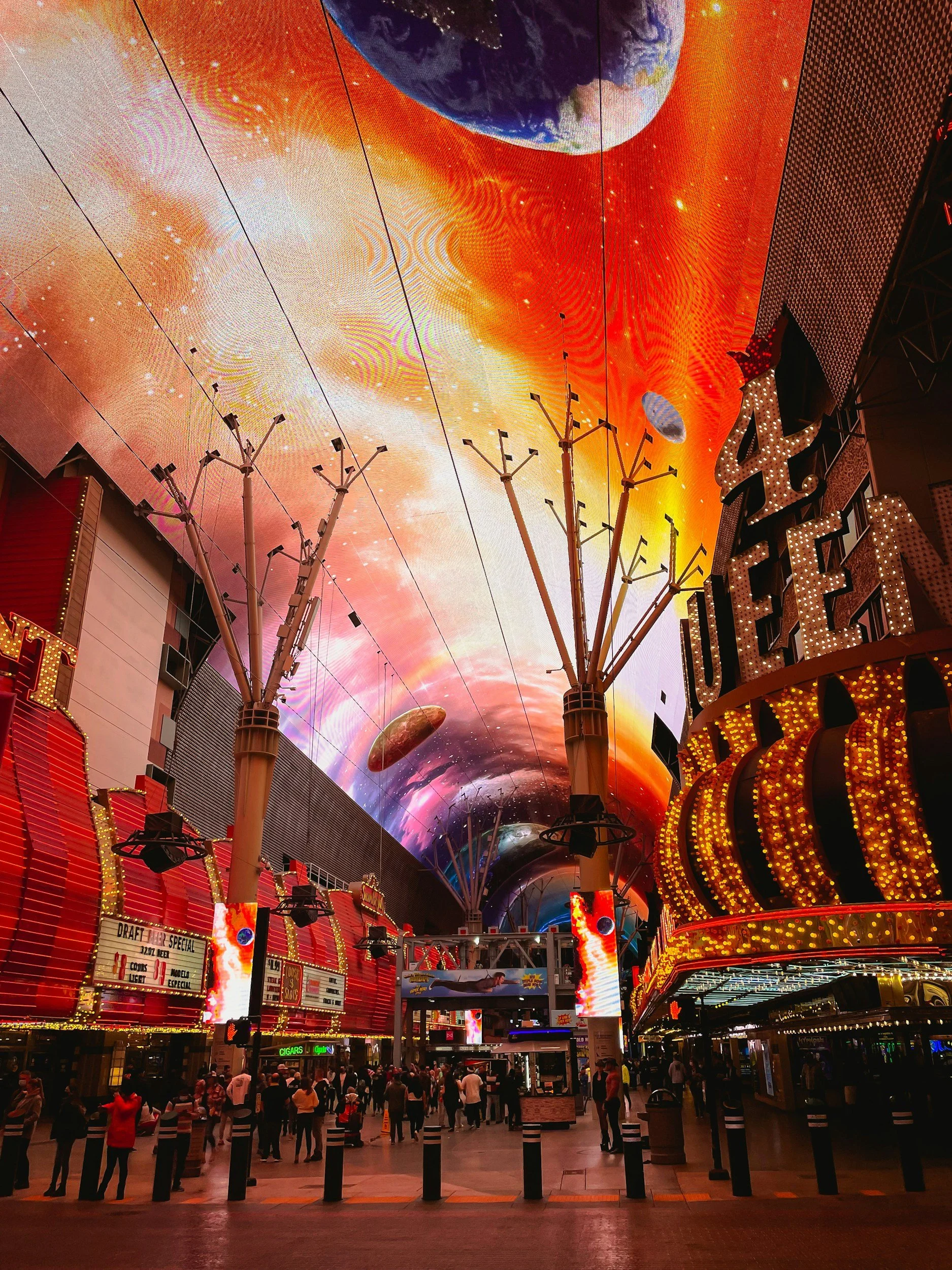 Bright lights and large digital screens inside a lively entertainment venue, with a crowd of people walking and standing in the foreground.