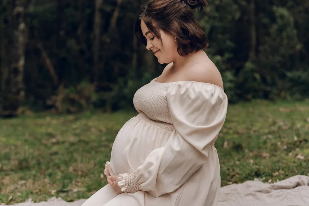 Pregnant woman sitting outdoors on a blanket, holding her belly and smiling gently.