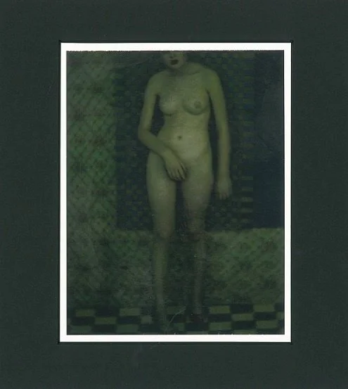 Nude woman standing in front of patterned fabric background, holding her chest with one hand, in a dimly lit room.