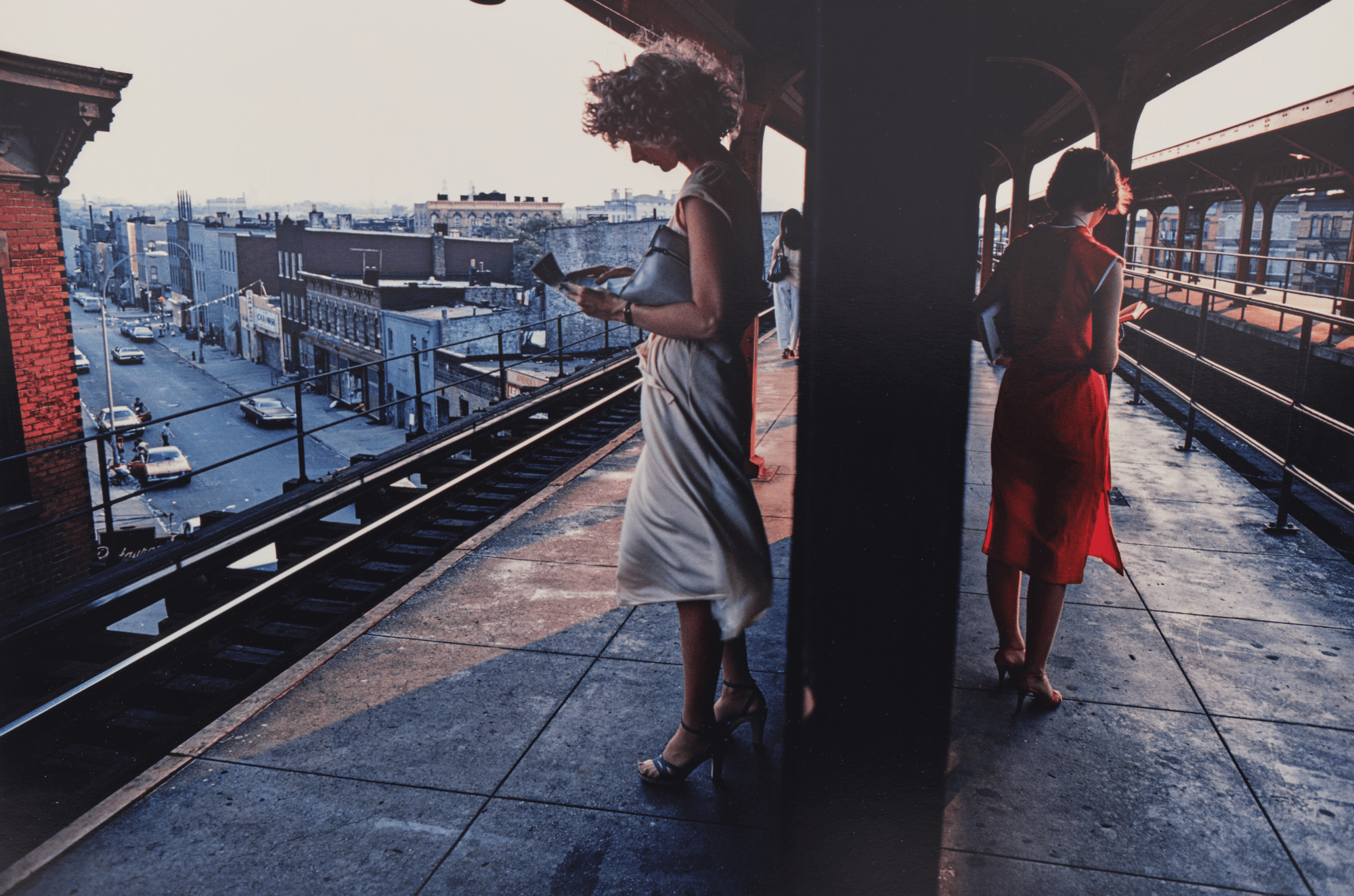 Bruce Davidson. Two women dressed in stylish dresses and high heels are standing on an elevated train platform, waiting near the edge and looking at their phones as the sunset casts a warm glow over the cityscape below.