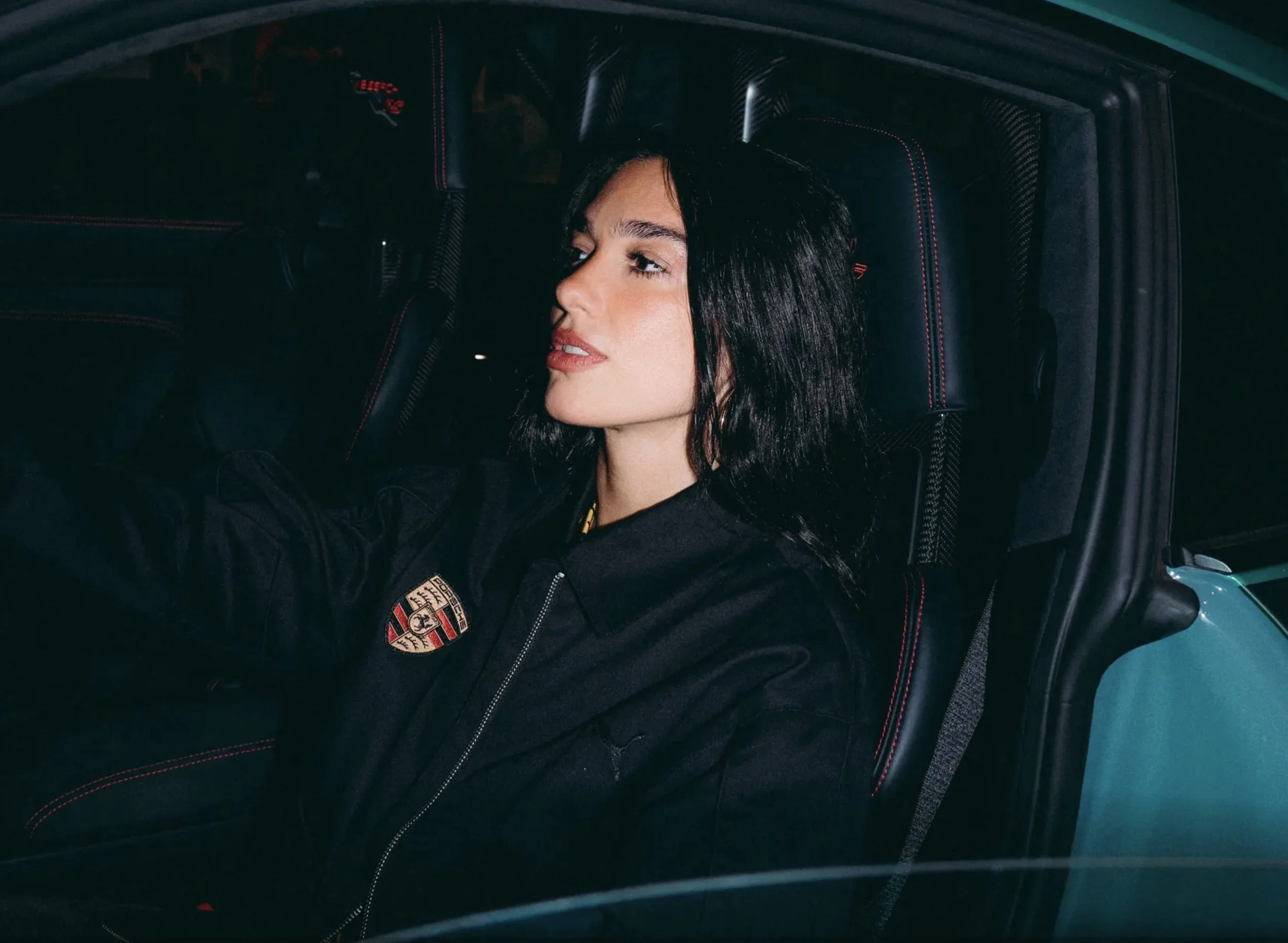 A young woman with black hair and red lipstick sitting in the driver's seat of a car, wearing a black jacket with a Porsche logo, looking to the side inside a modern car interior.