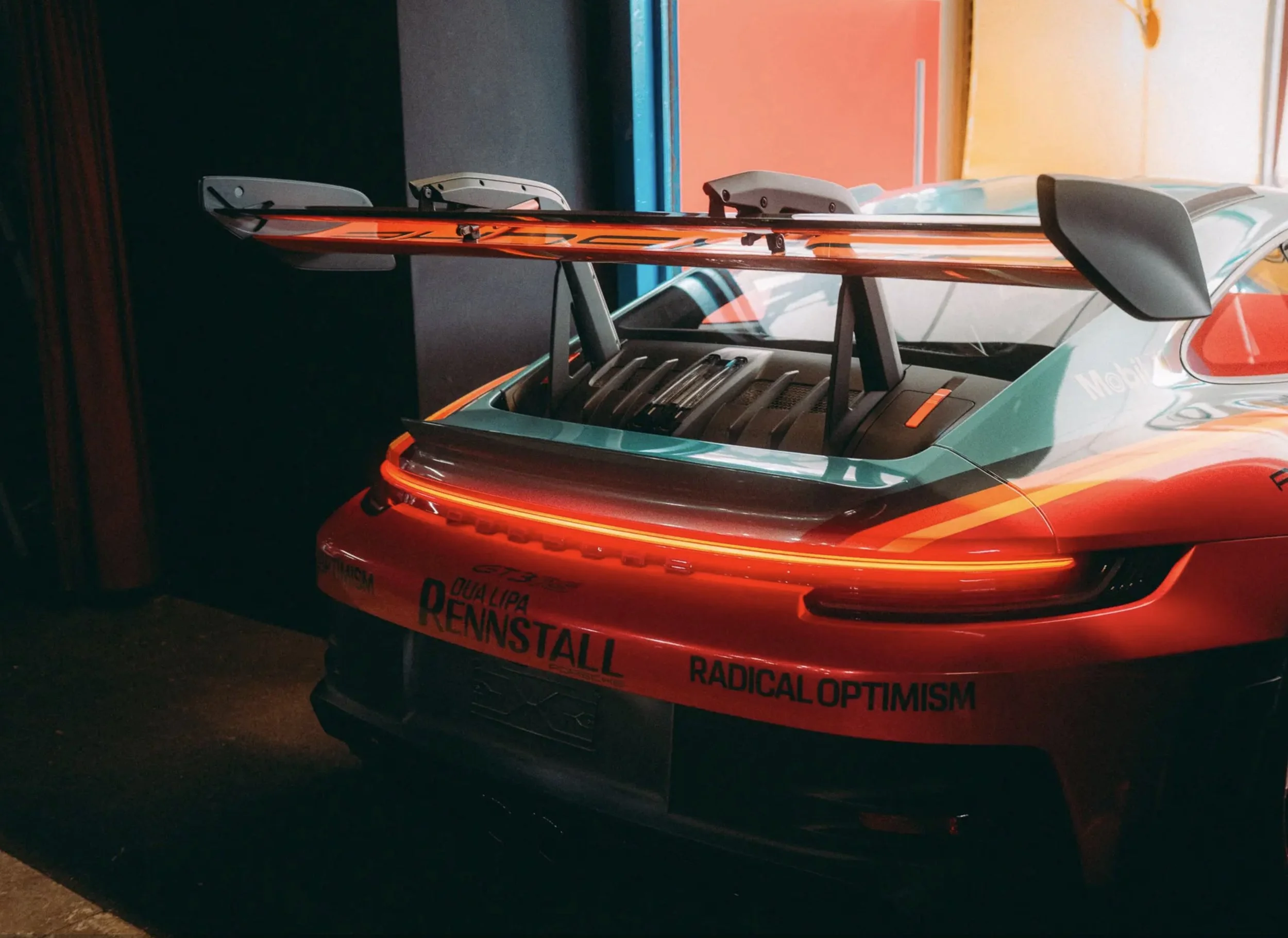 The rear end of a bright orange sports car with a large rear wing, parked indoors under warm lighting. The car has various logos and text on the bumper, including 'QUA LIPA RENNSTALL' and 'RADICAL OPTIMISM.