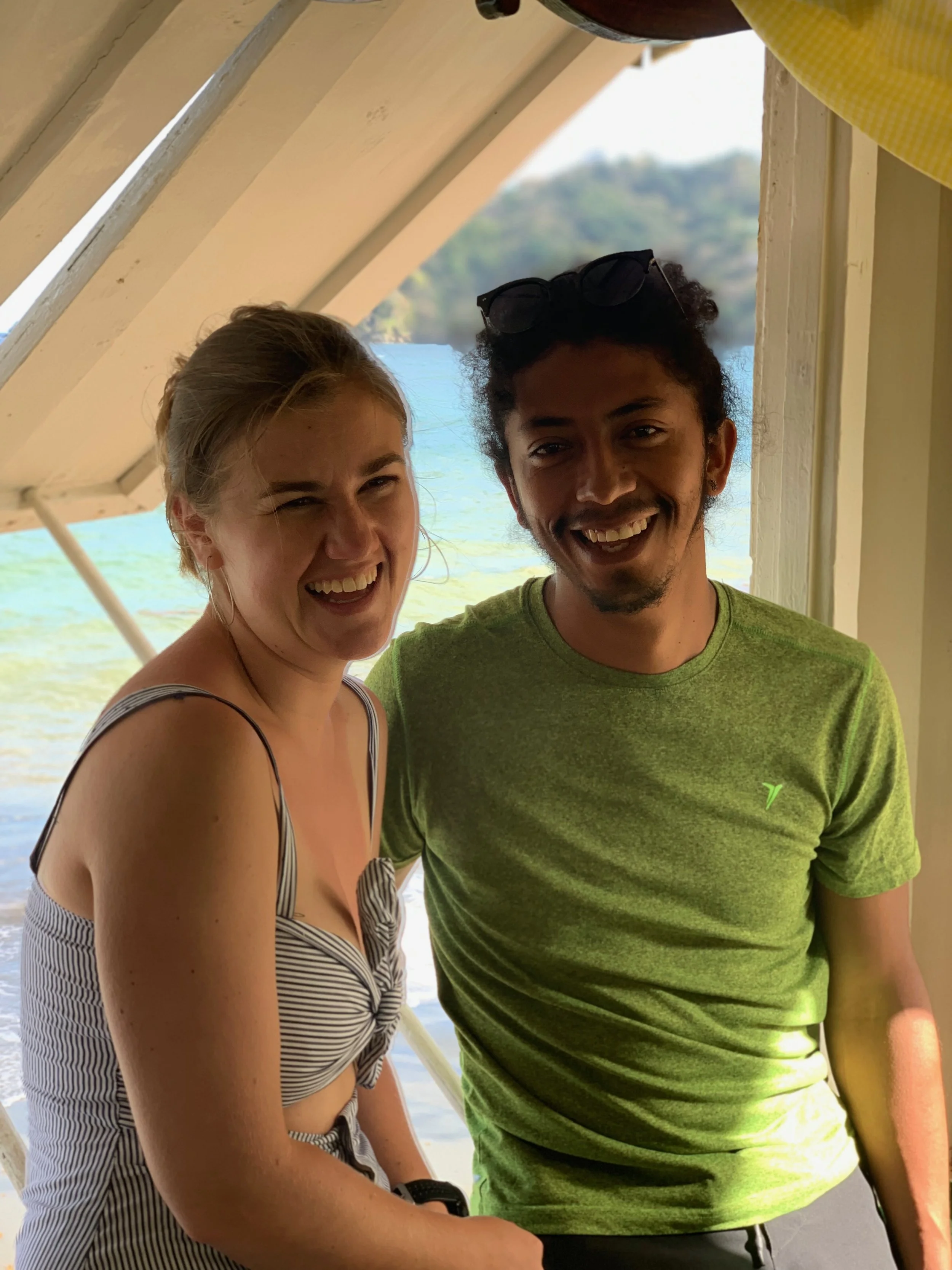 Close-up photo of a young woman and a young man smiling outdoors near water, with a lush background. The woman has blonde hair, wears a striped sundress, and the man has curly hair, sunglasses on his head, and wears a green T-shirt.