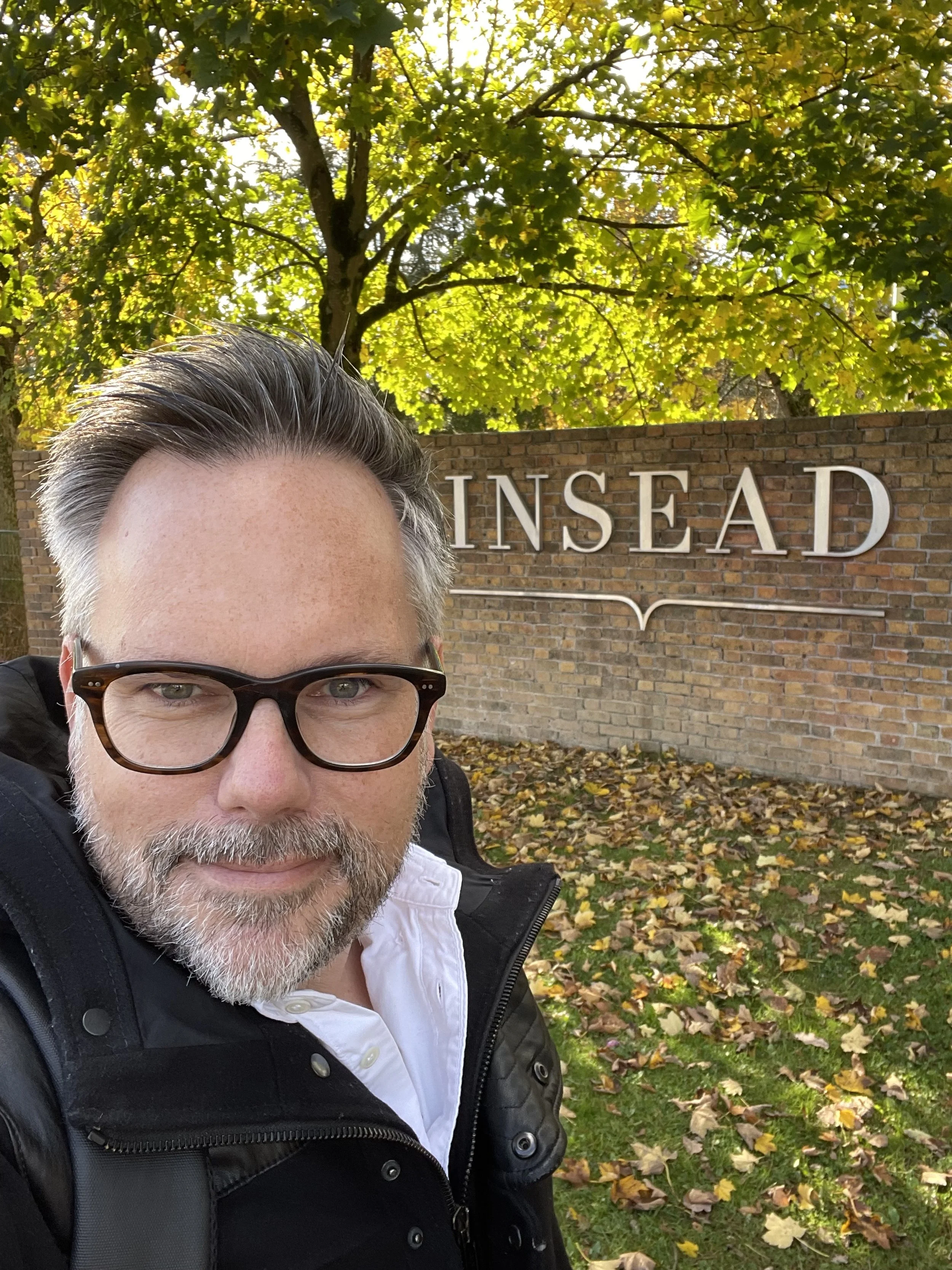 Robin Karakashmit Brille und grauem Bart vor einem Zaun mit der Universität 'INSEAD' steht, umgeben von Bäumen im Herbst und feiert seinen Abschluss und Zertifikat im Executive Coaching.