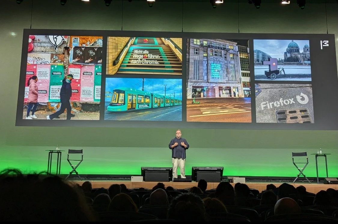 Eine Präsentation auf einer großen Leinwand zeigt verschiedene Werbemotive von einer Firefox Kampagne. Robin Karakash steht vor Publikum auf der Bühne und präsentiert diese.