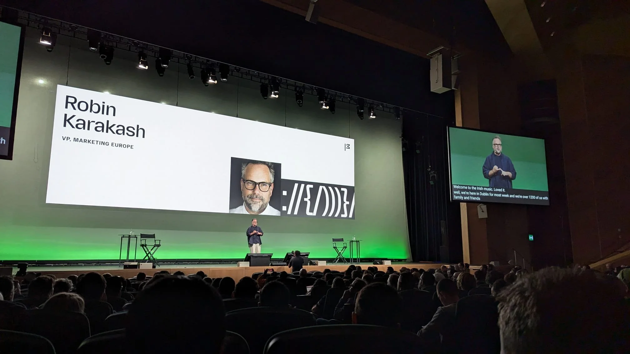 Vortrag auf einer großen Bühne mit einer Präsentationsfolie, die den Namen Robin Karakash und seine Position als VP Marketing Europa zeigt, sowie ein Portraitfoto. Es sitzen viele Leute im Publikum, außerdem gibt es einen kleinen Bildschirm rechts mit einer Sign Language Translator.