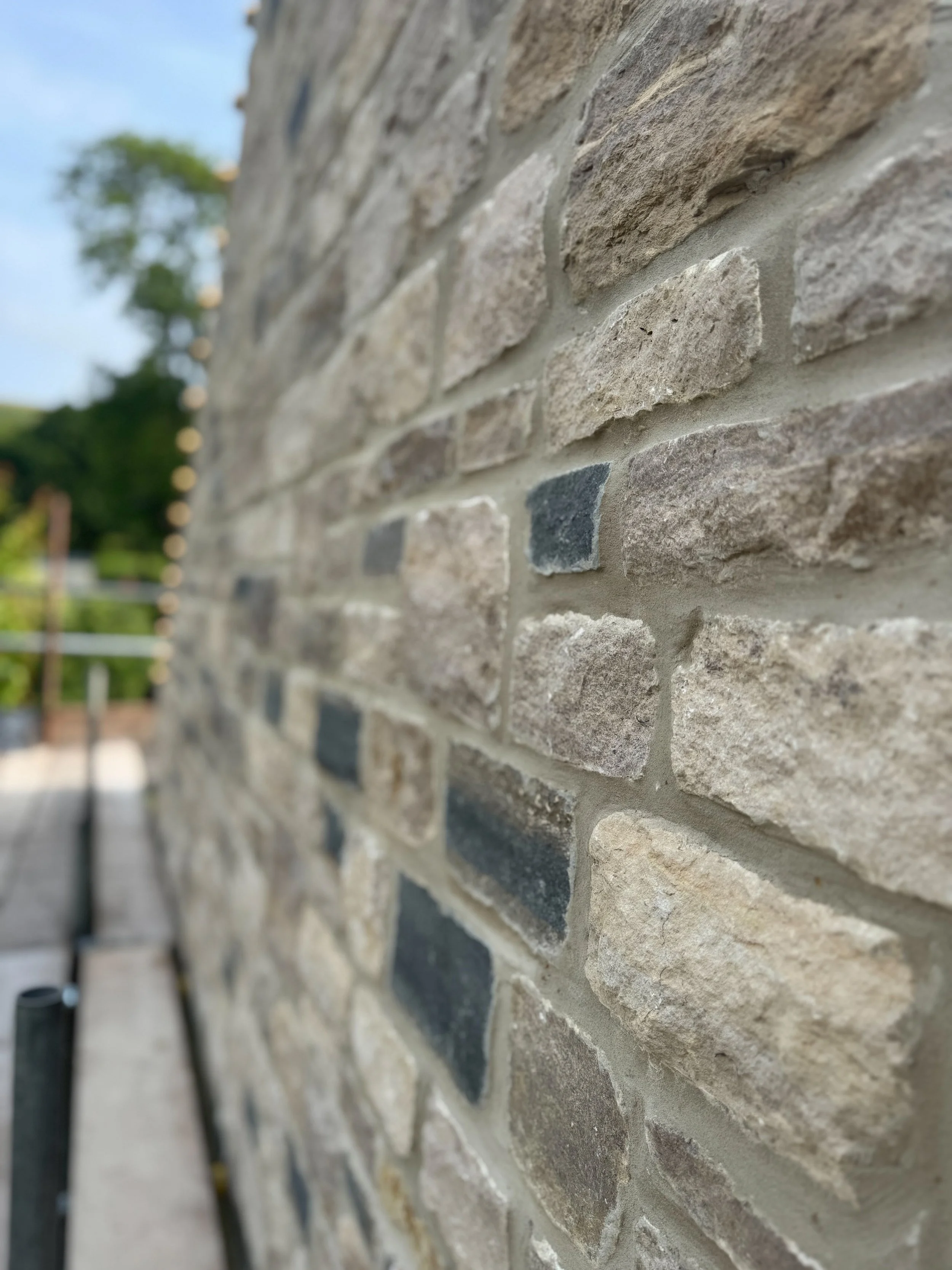 Close-up of a textured stone brick wall with light gray mortar, featuring various shades of beige, brown, and dark gray stones. In the background, blurred greenery and a clear sky are visible.