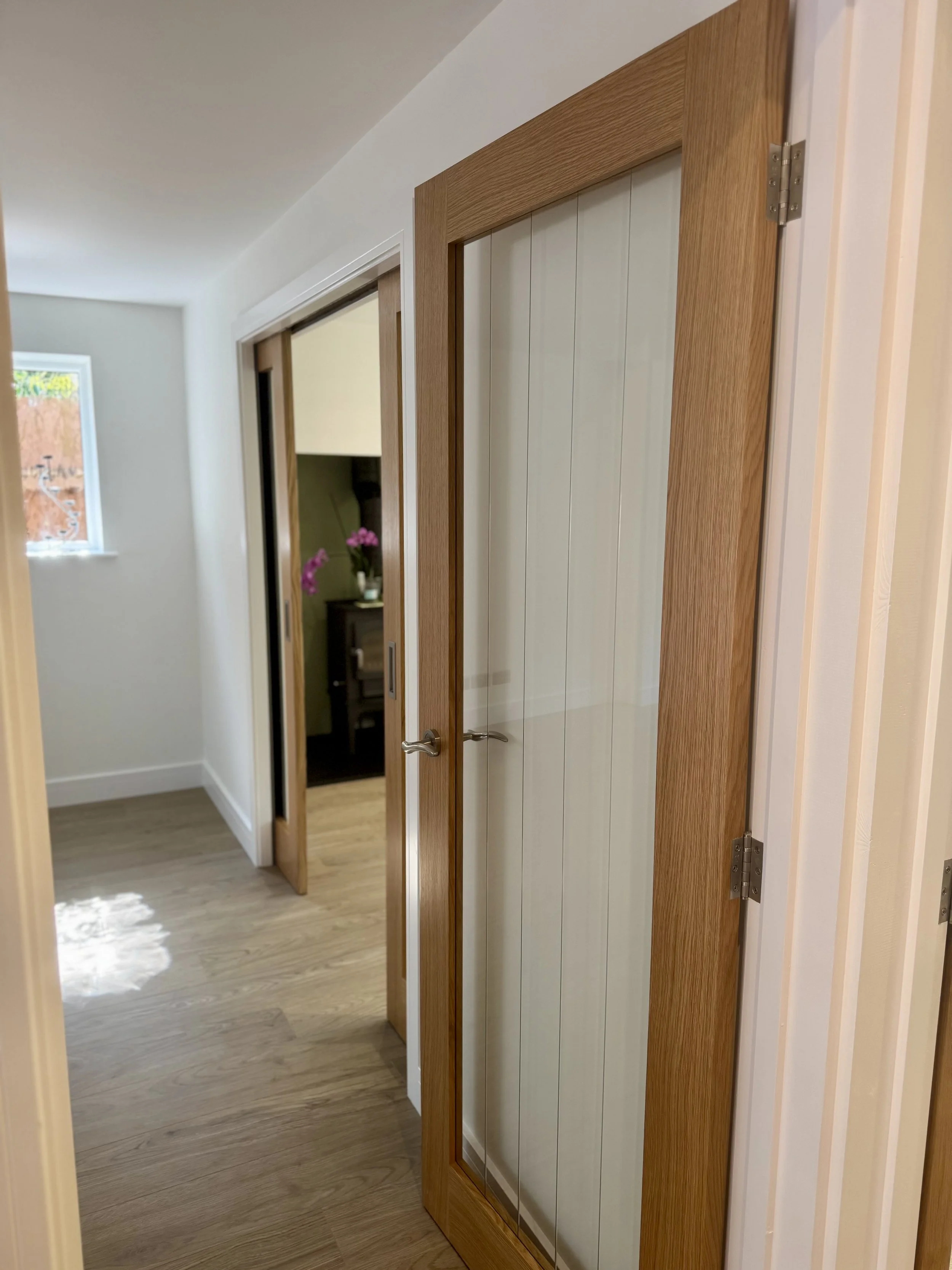 Interior view of a hallway in a house, showing a wooden framed glass door, two open closet doors, and a room with a dark cabinet and pink flowers in a vase.