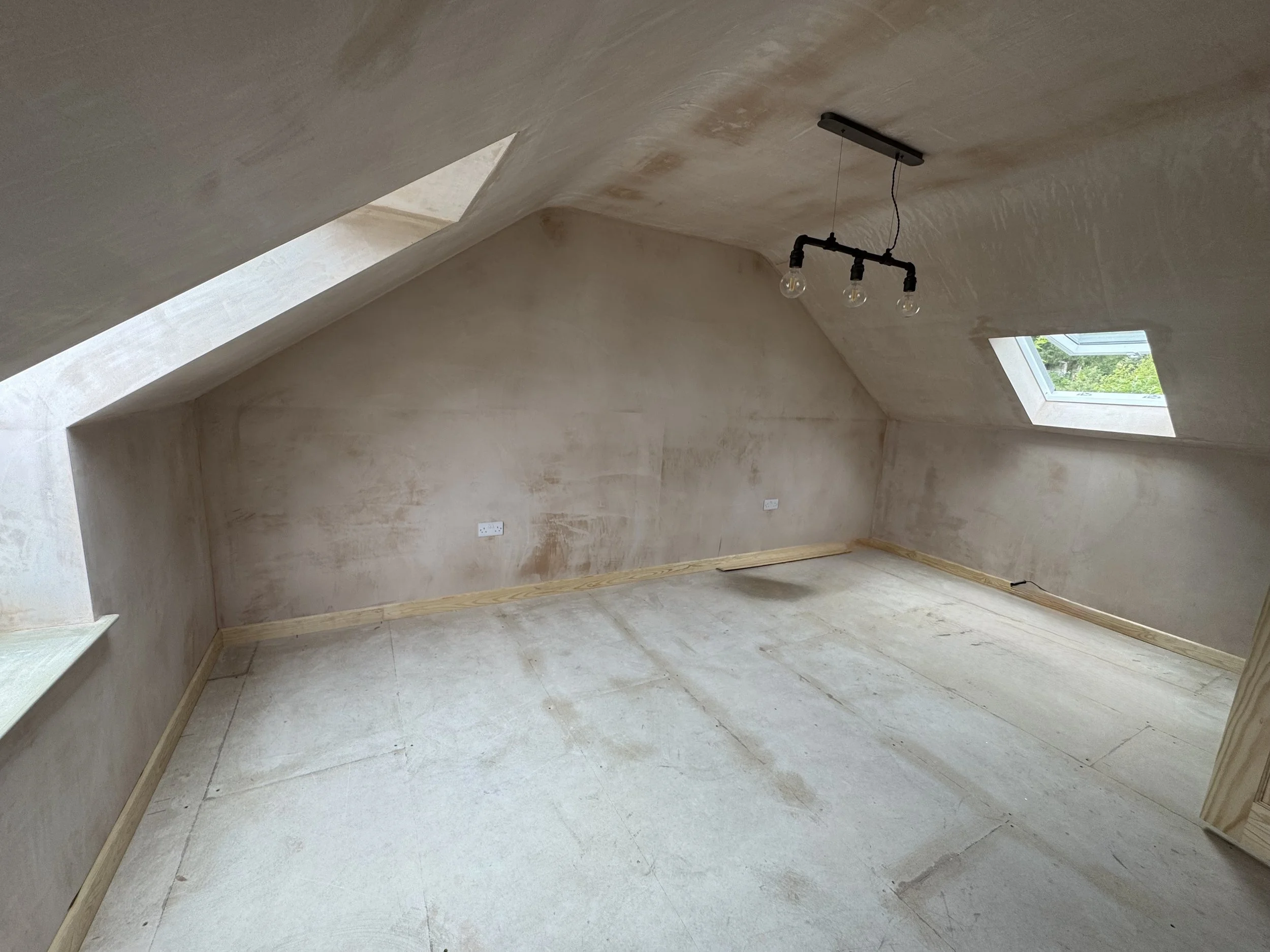 Empty attic room with sloped ceiling and two skylight windows, beige walls, and exposed light fixture with three bulbs hanging from the ceiling.