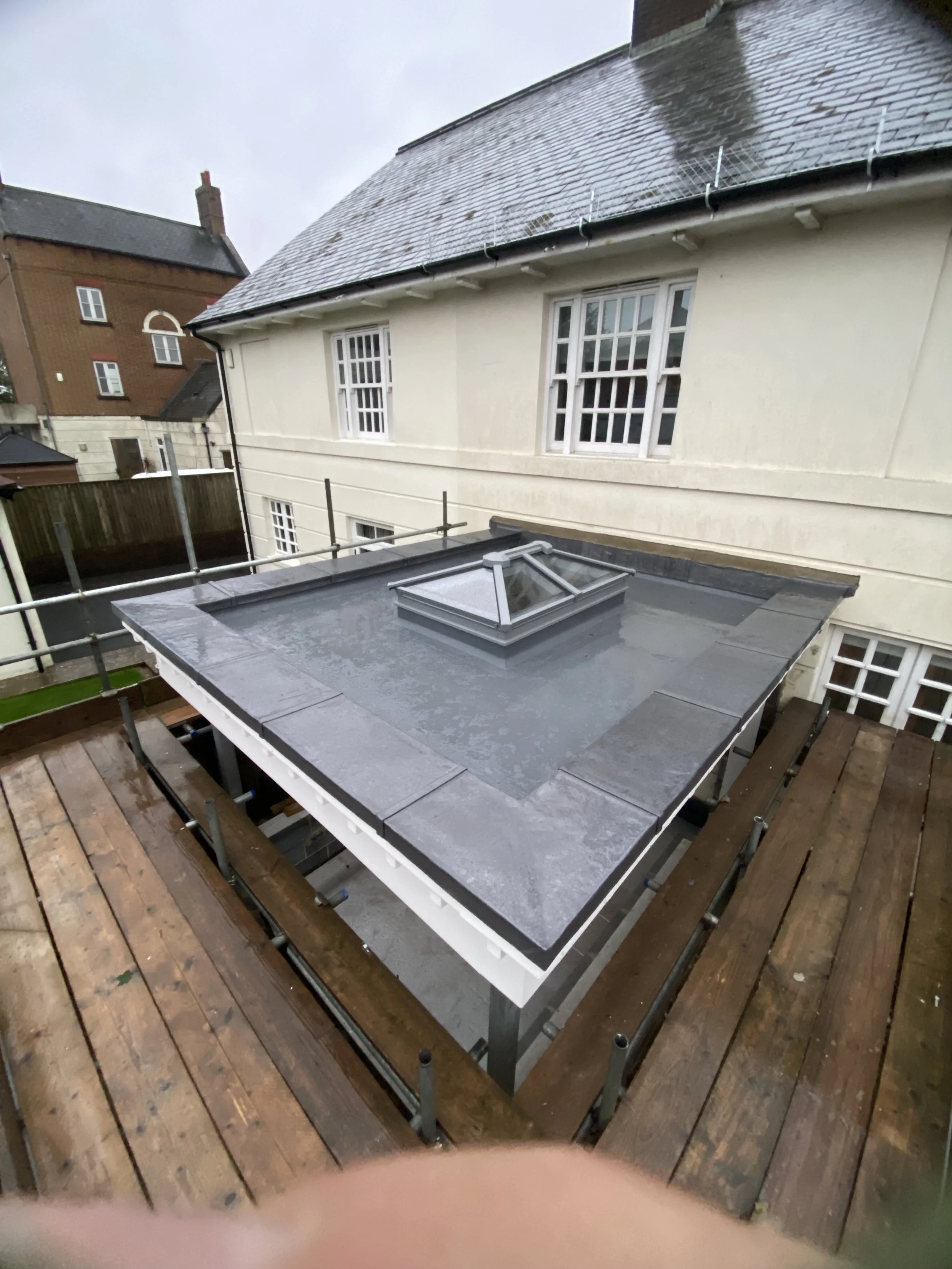 View of a flat roof with a skylight, surrounded by building walls, with some weathering and a wooden deck in the foreground, under an overcast sky.