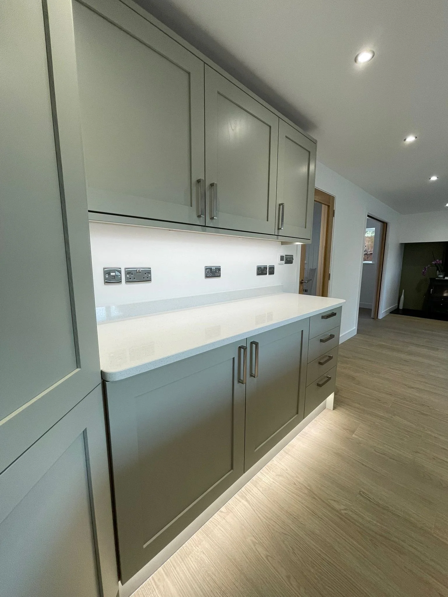 Modern kitchen with light gray cabinets, white countertop, and multiple electrical outlets along the backsplash, with wooden flooring and recessed ceiling lights.