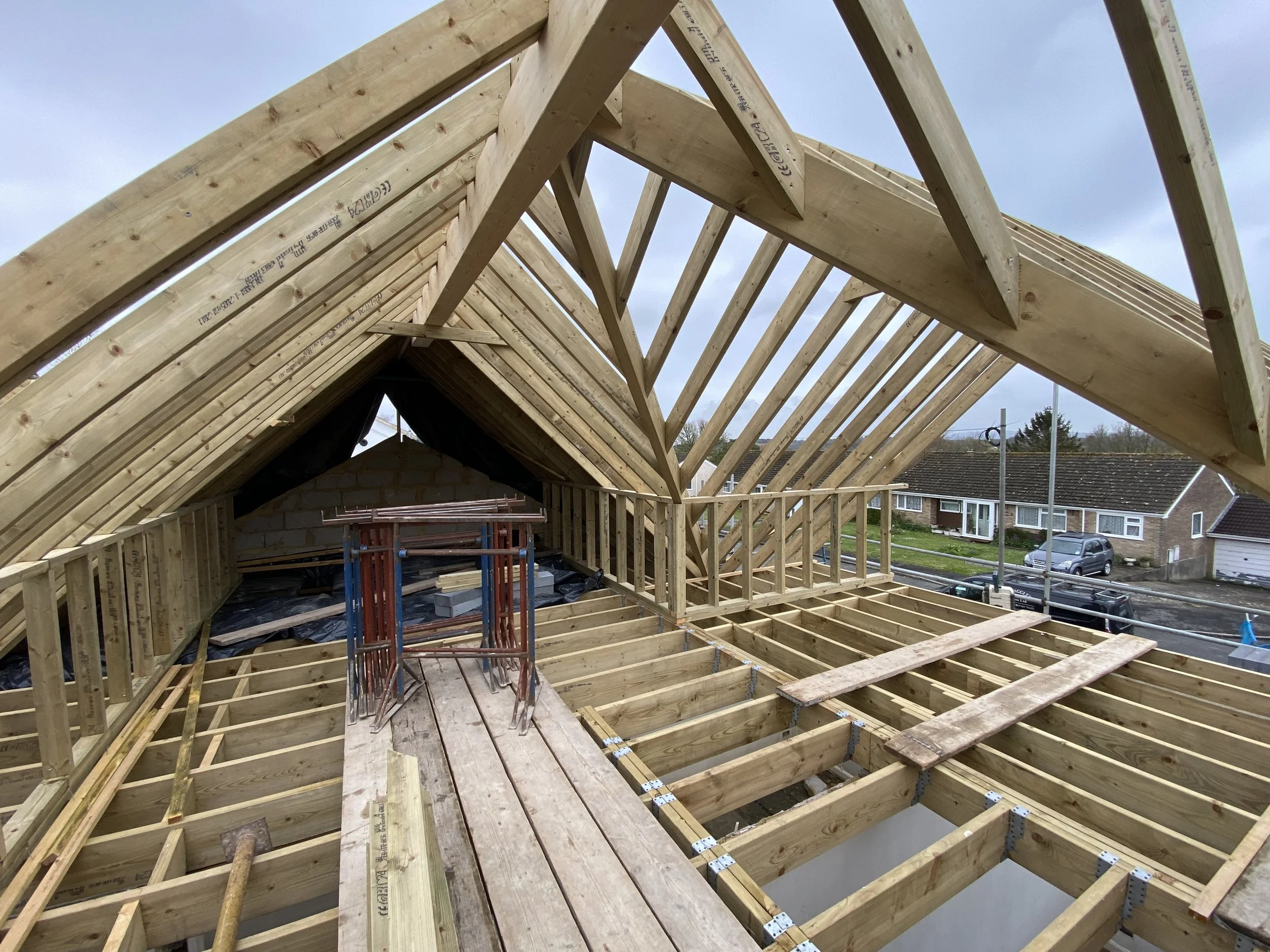 Construction site with wooden framing for a house roof and floor, scaffolding, and nearby neighborhood houses visible in the background.