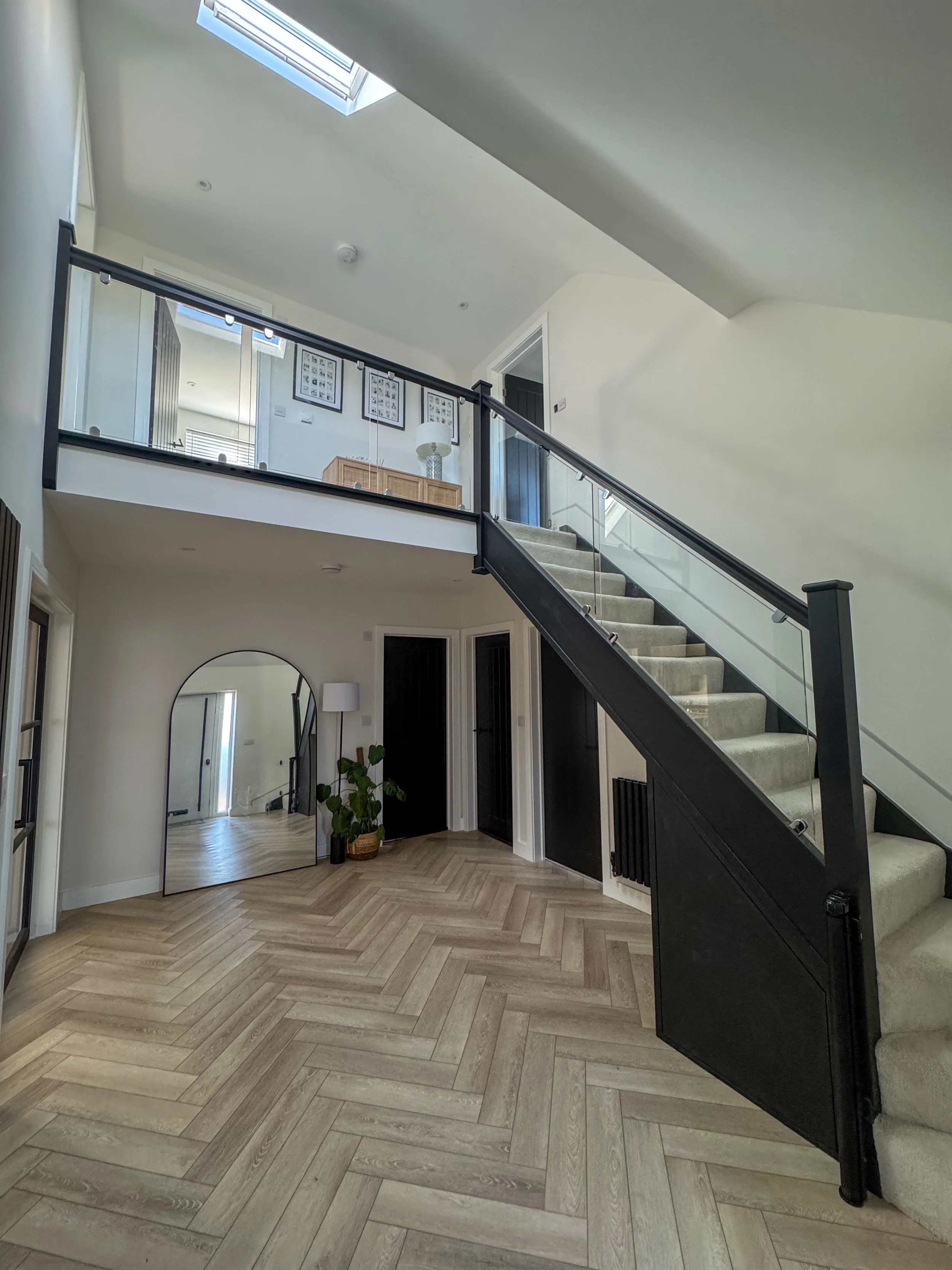 Modern interior with a staircase leading to a second-floor balcony, a large mirror on the floor, potted plant, and skylights illuminating hardwood herringbone floor.
