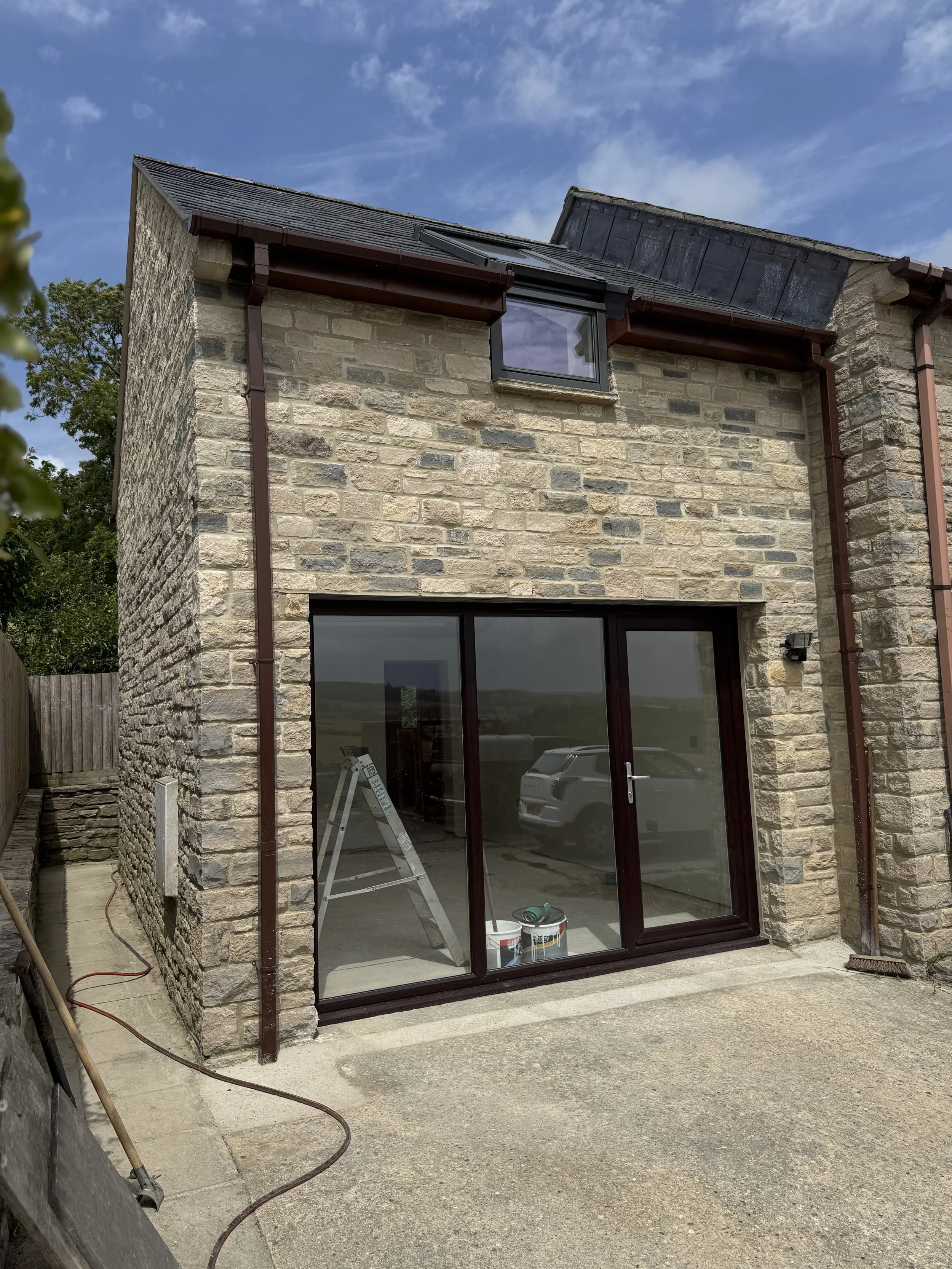 Newly constructed stone house with large glass sliding door and small roof window, outdoor construction tools and buckets visible, partly cloudy sky in background.