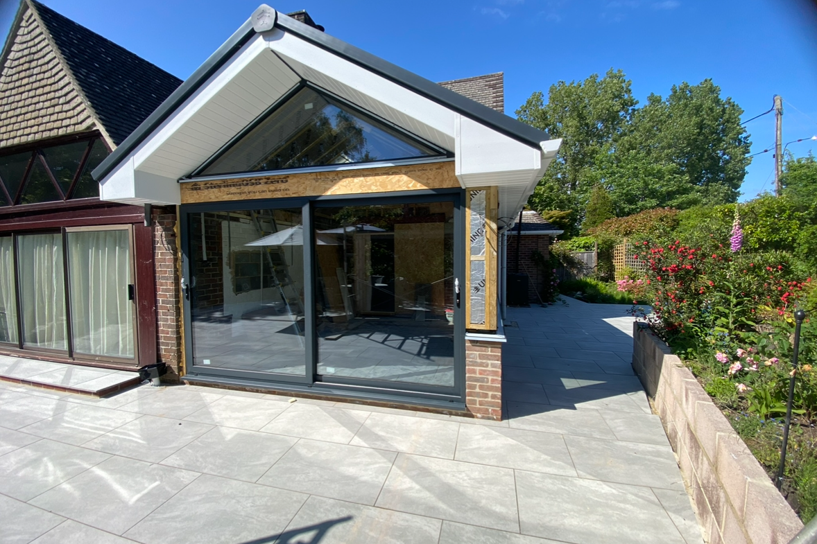 A house with a newly constructed extension featuring large glass sliding doors, a sloped roof with a triangular window, and outdoor tiled patio area, with a garden and trees in the background.