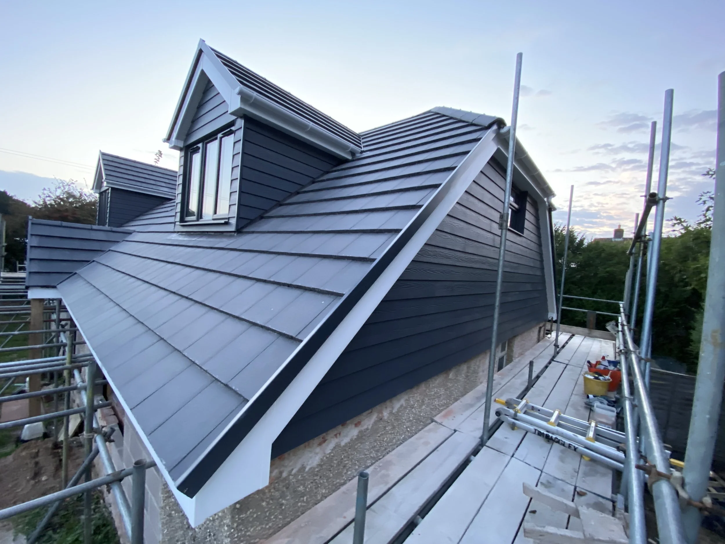 A house under construction with a newly installed dark blue siding and a metal roof. Scaffolding surrounds the house, and tools are visible on the platform.