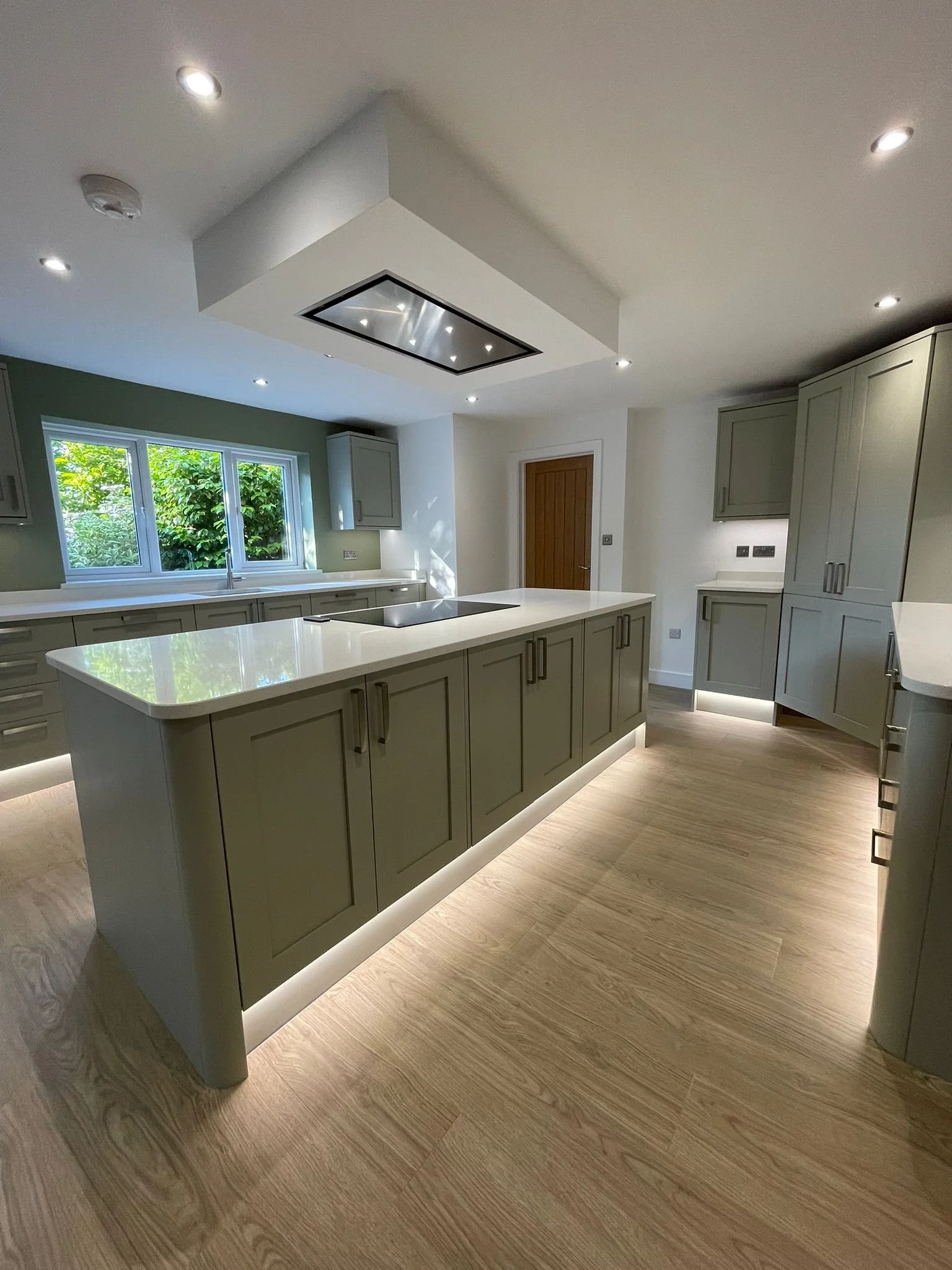 Modern kitchen with beige cabinetry, a central island with a smooth countertop, wooden flooring, a window with green foliage outside, and recessed lighting.
