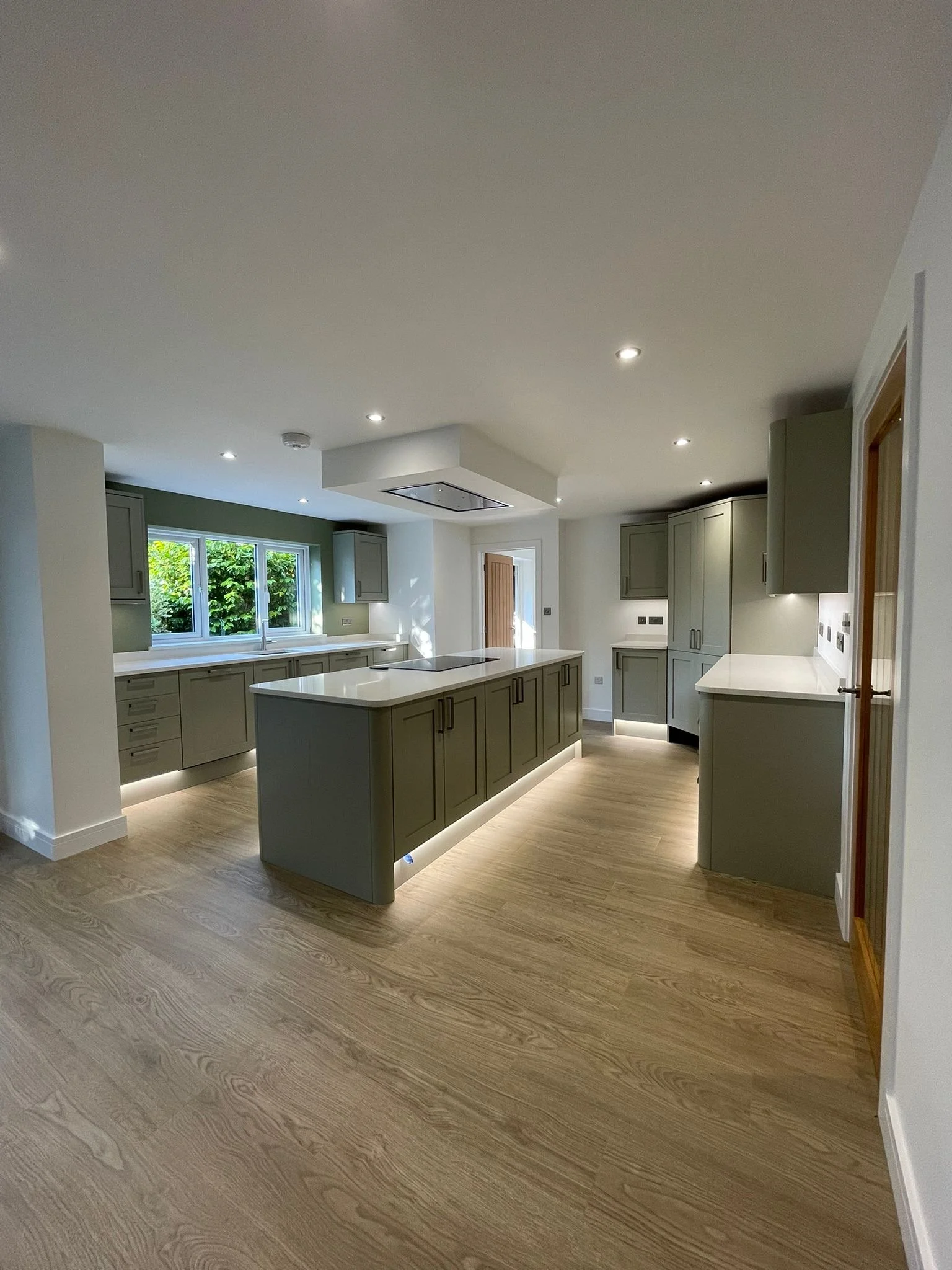 Modern kitchen with gray cabinets, white countertops, a central island, wooden flooring, and green foliage visible through a window.