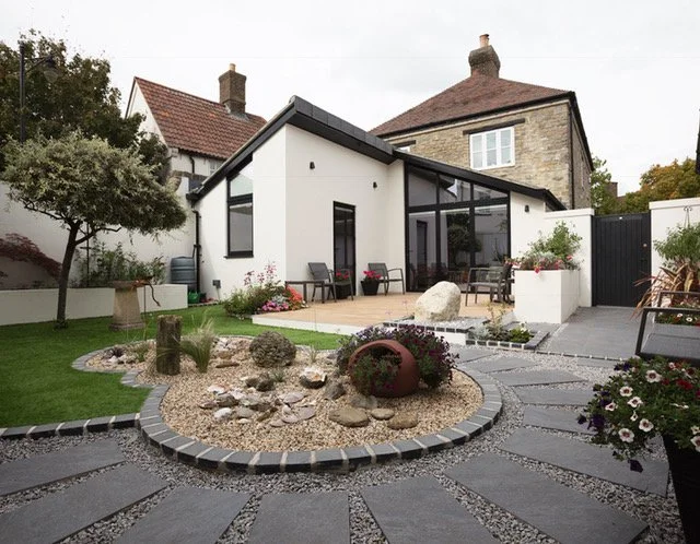 Modern backyard with flagstone pathway, rock garden, and seating area, adjacent to a white house with large glass windows