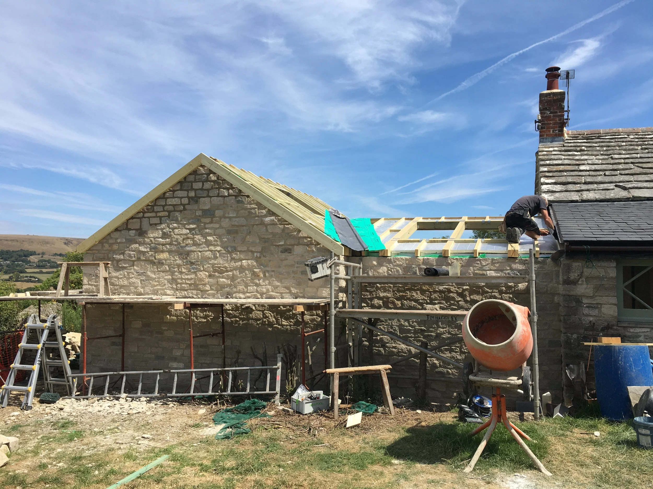 Construction workers building a roof on a stone house with scaffolding, ladders, and construction tools around the site, against a blue sky with wispy clouds.