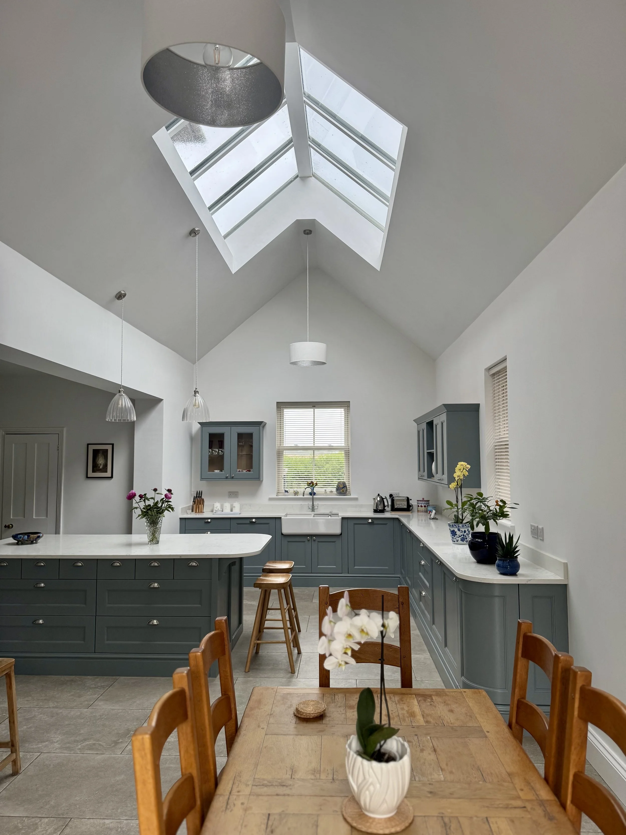 Modern kitchen with gray cabinets, a skylight ceiling, a window above the sink, wooden chairs, a wooden dining table with a potted orchid plant, and hanging pendant lights.