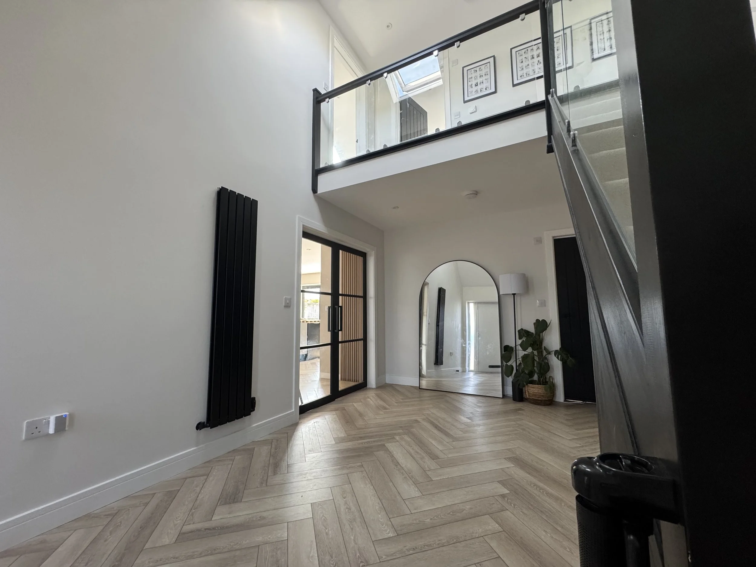 Modern interior with white walls, wooden chevron flooring, a large floor mirror, a black vertical radiator, a sliding glass door, potted plant, standing lamp, and a mezzanine with glass railing.