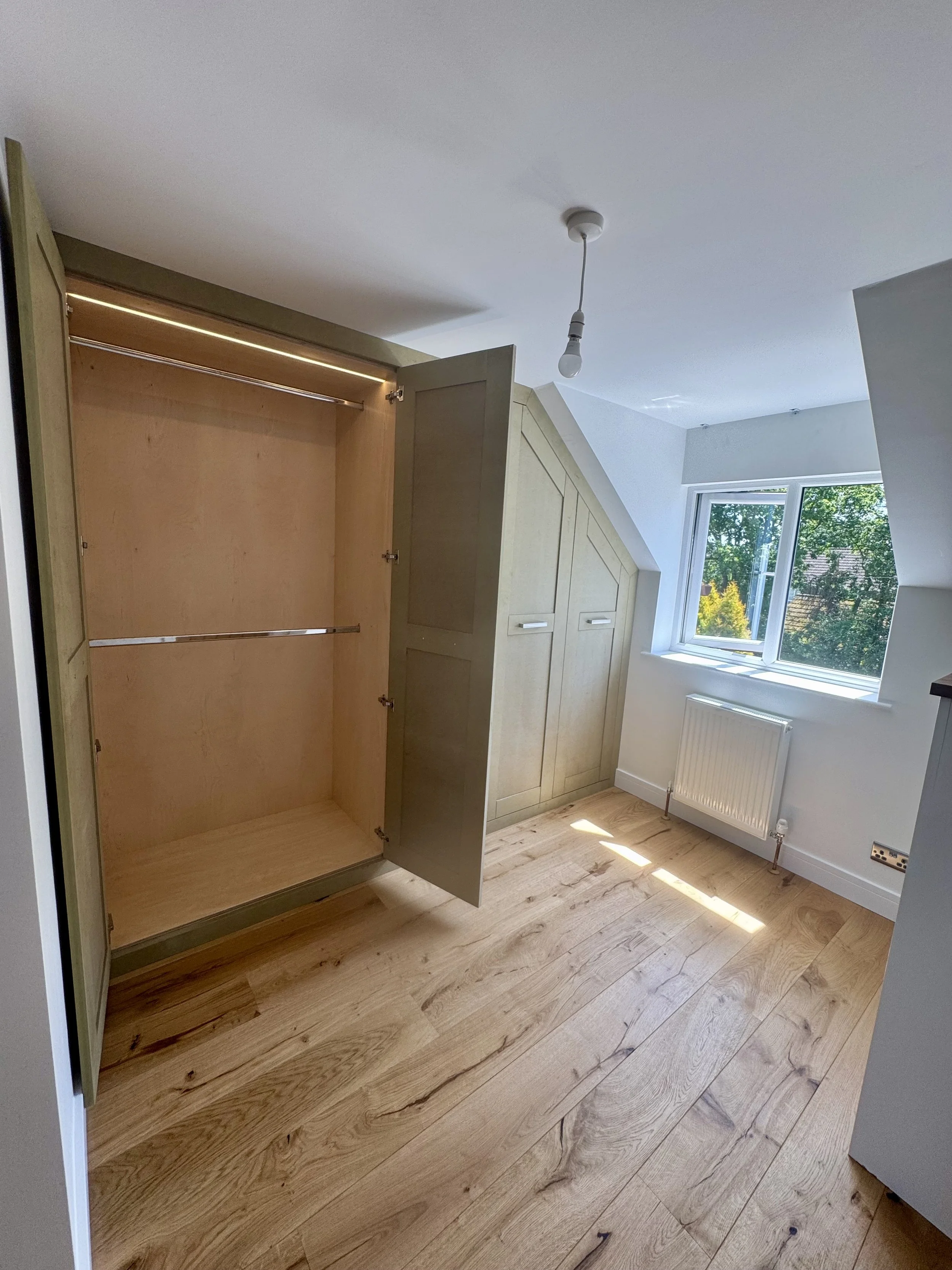 Empty bedroom with built-in wardrobe, window with greenery outside, and wooden floor.