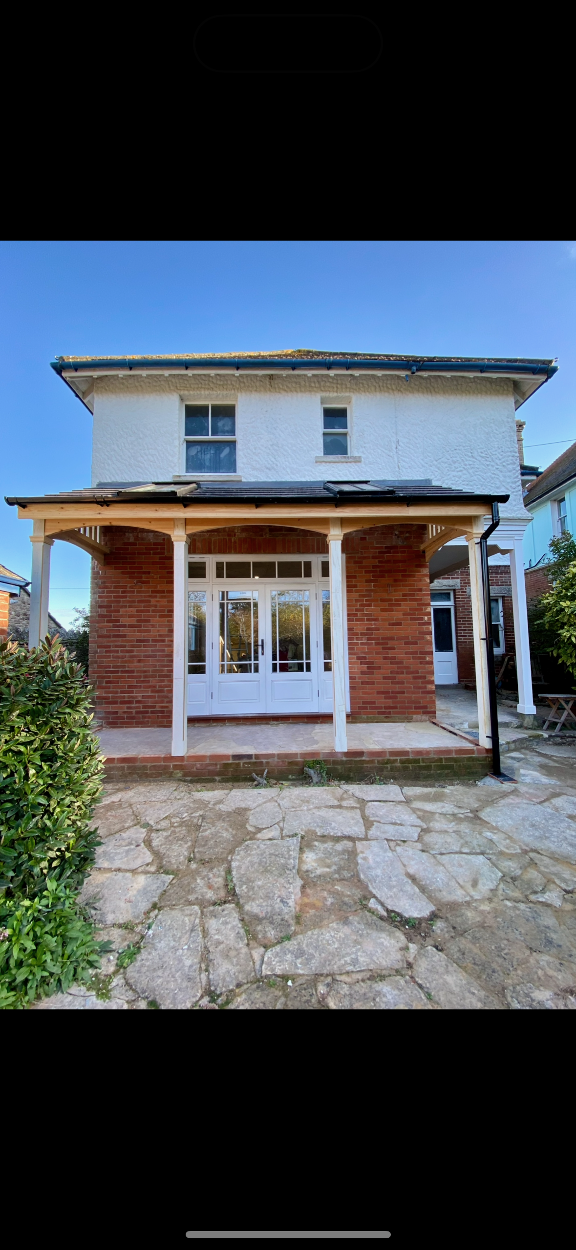 A two-story house with a brick and white exterior, a covered porch with a wooden roof structure, glass front doors, and a stone patio, under a clear blue sky.