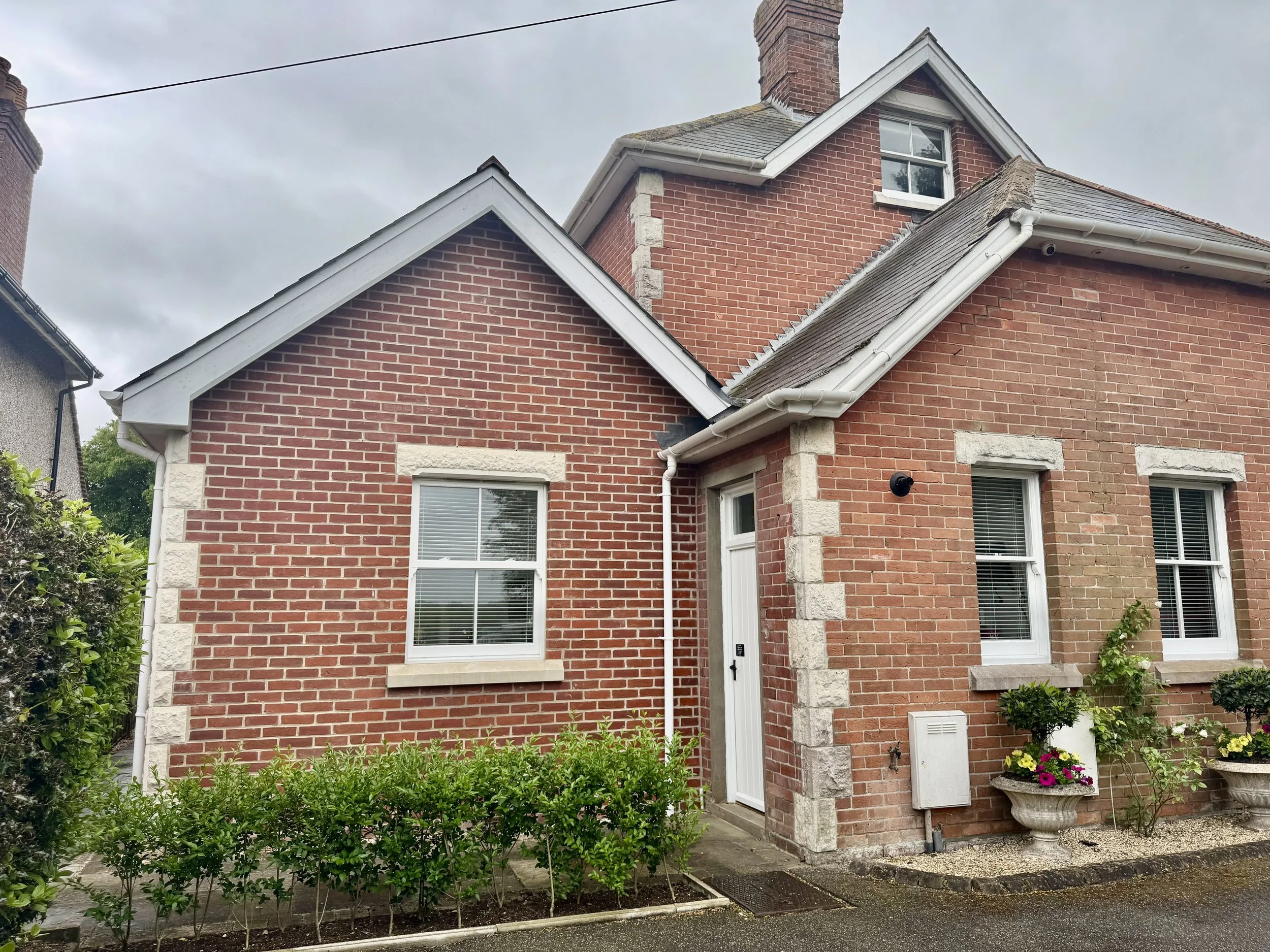 Brick house with white-framed windows, a white door, potted plants, and a bush in front under an overcast sky.