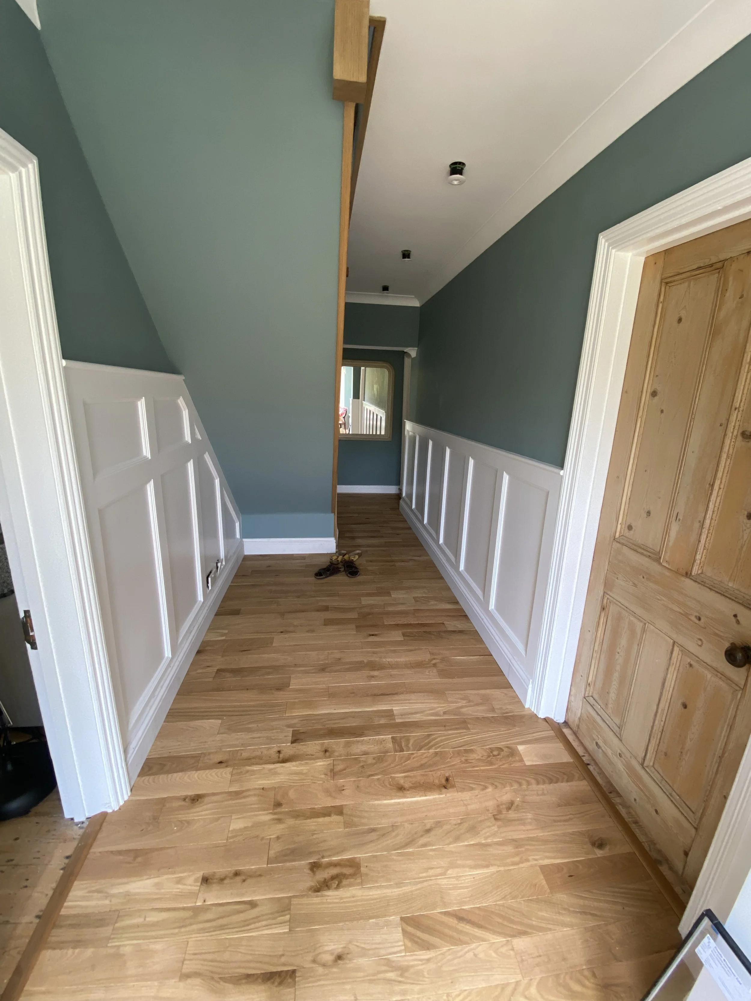 Hallway with wooden flooring, green upper walls, white wainscoting, and a wooden door on the right. Freshly painted walls and new flooring.