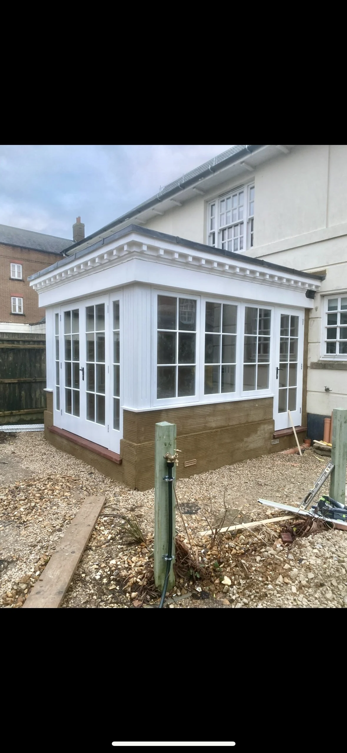 Newly built white conservatory or sunroom attached to a house, with large glass windows and double doors, on a gravel yard, under construction.