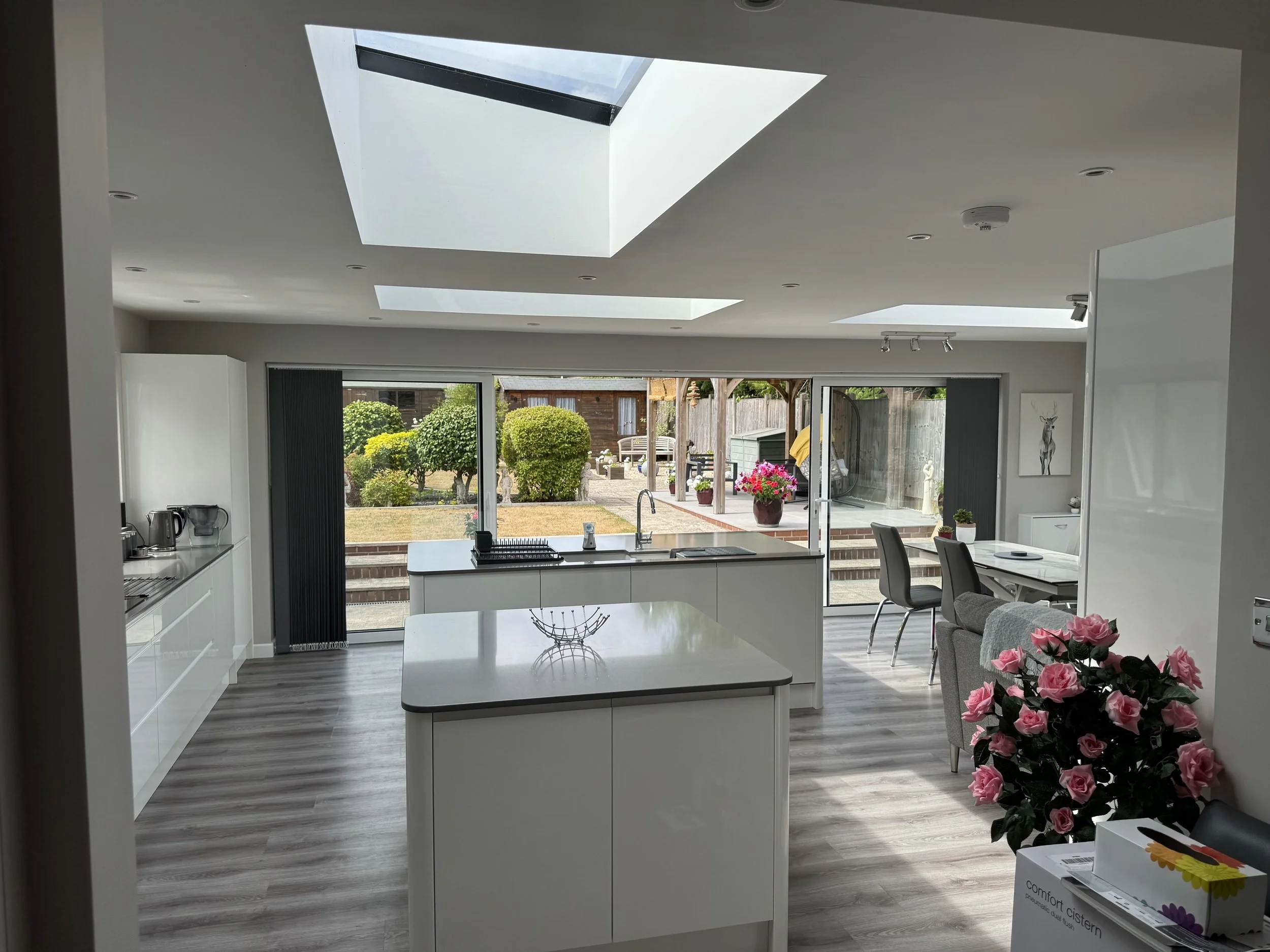 Modern kitchen with white cabinets, island, skylights, and sliding glass doors overlooking a garden with bushes and potted flowers.
