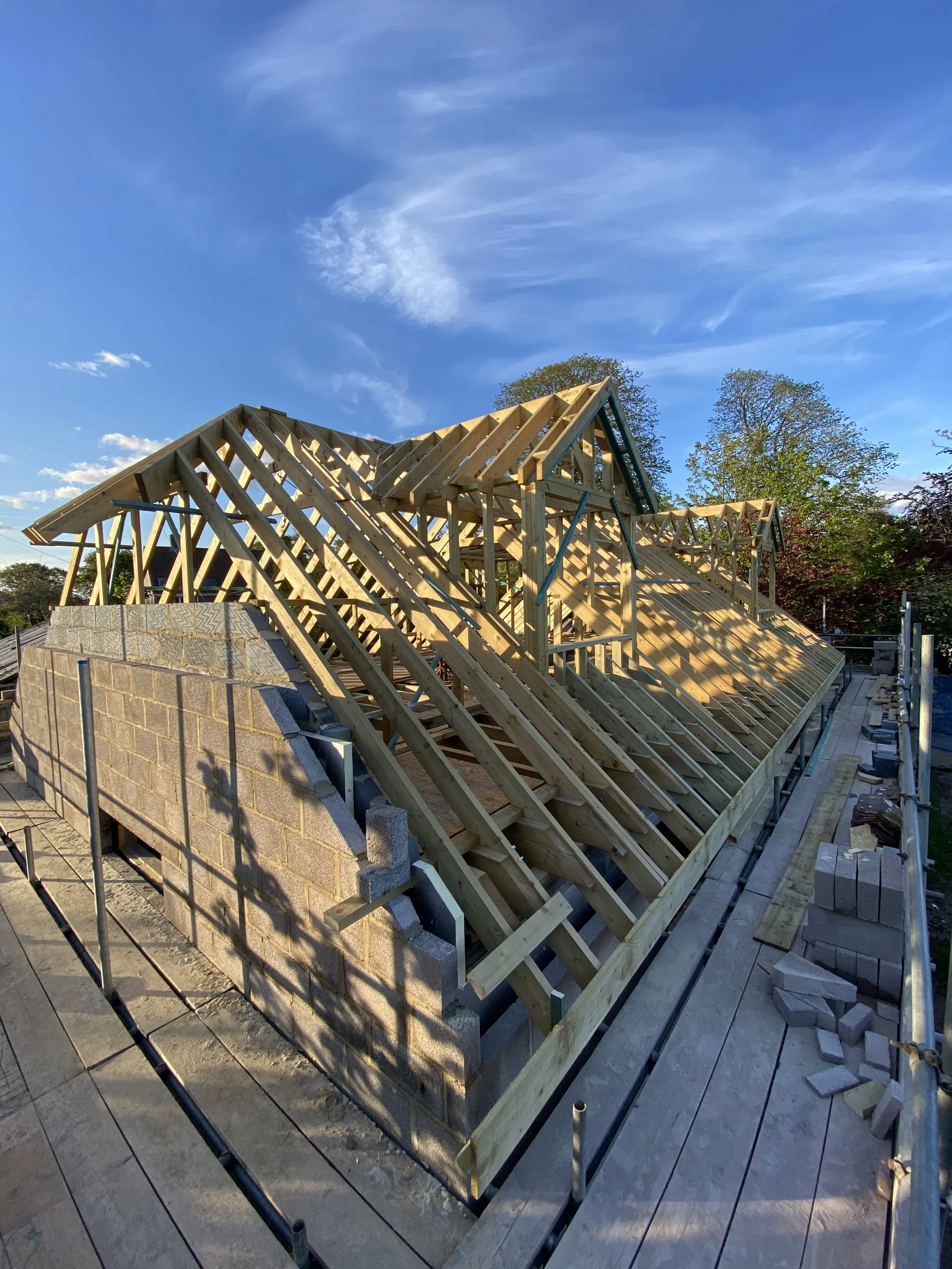 Wooden framework for a house roof under construction, with a brick wall at one side, and trees and a sky with clouds in the background.