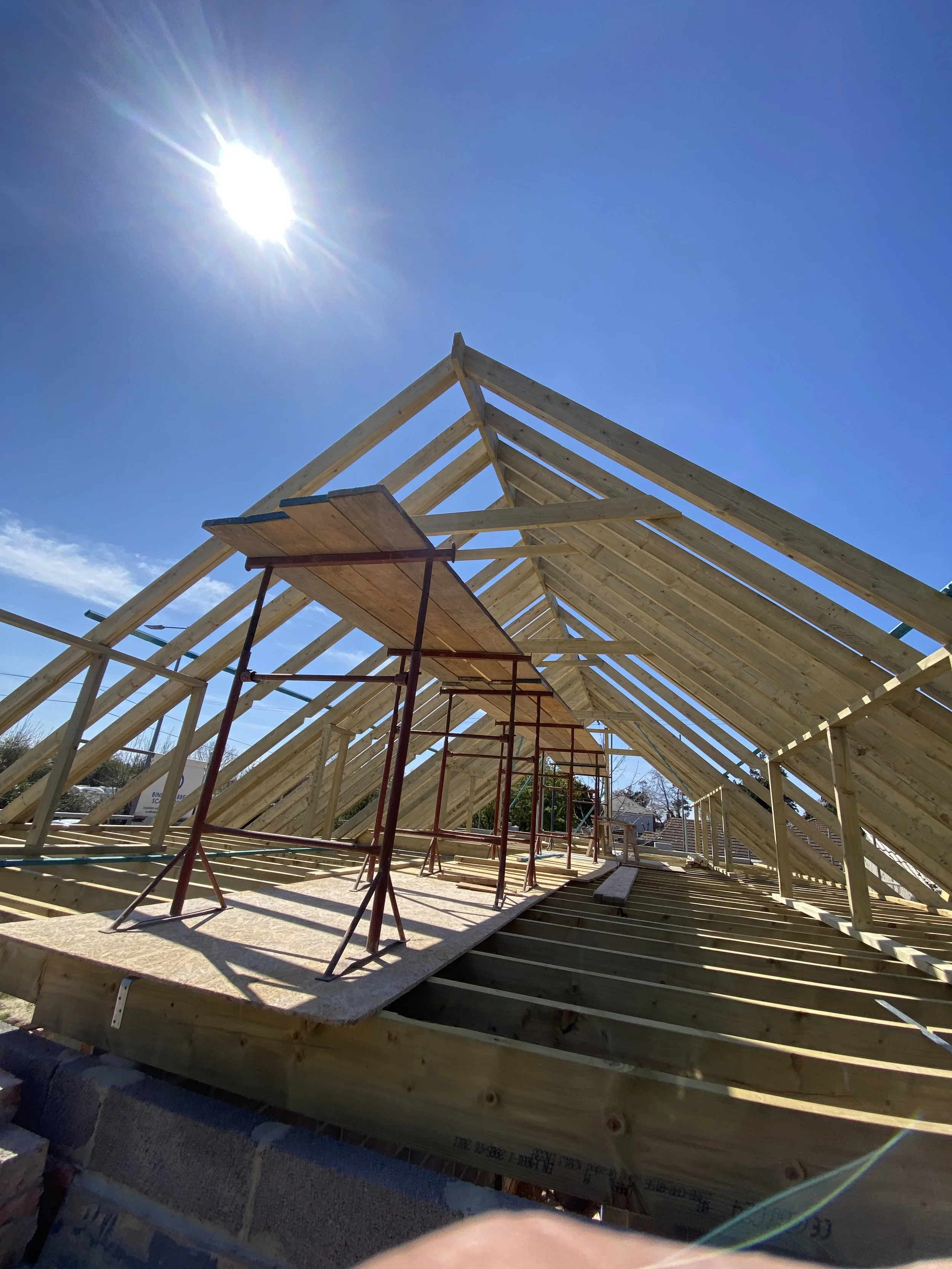 Construction site with a wooden roof framework and scaffolding, under a bright sun and a clear blue sky.