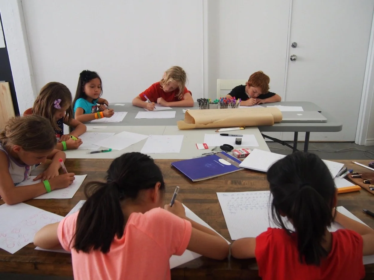 Group of children sitting at tables, writing on sheets of paper during an art or homework activity.