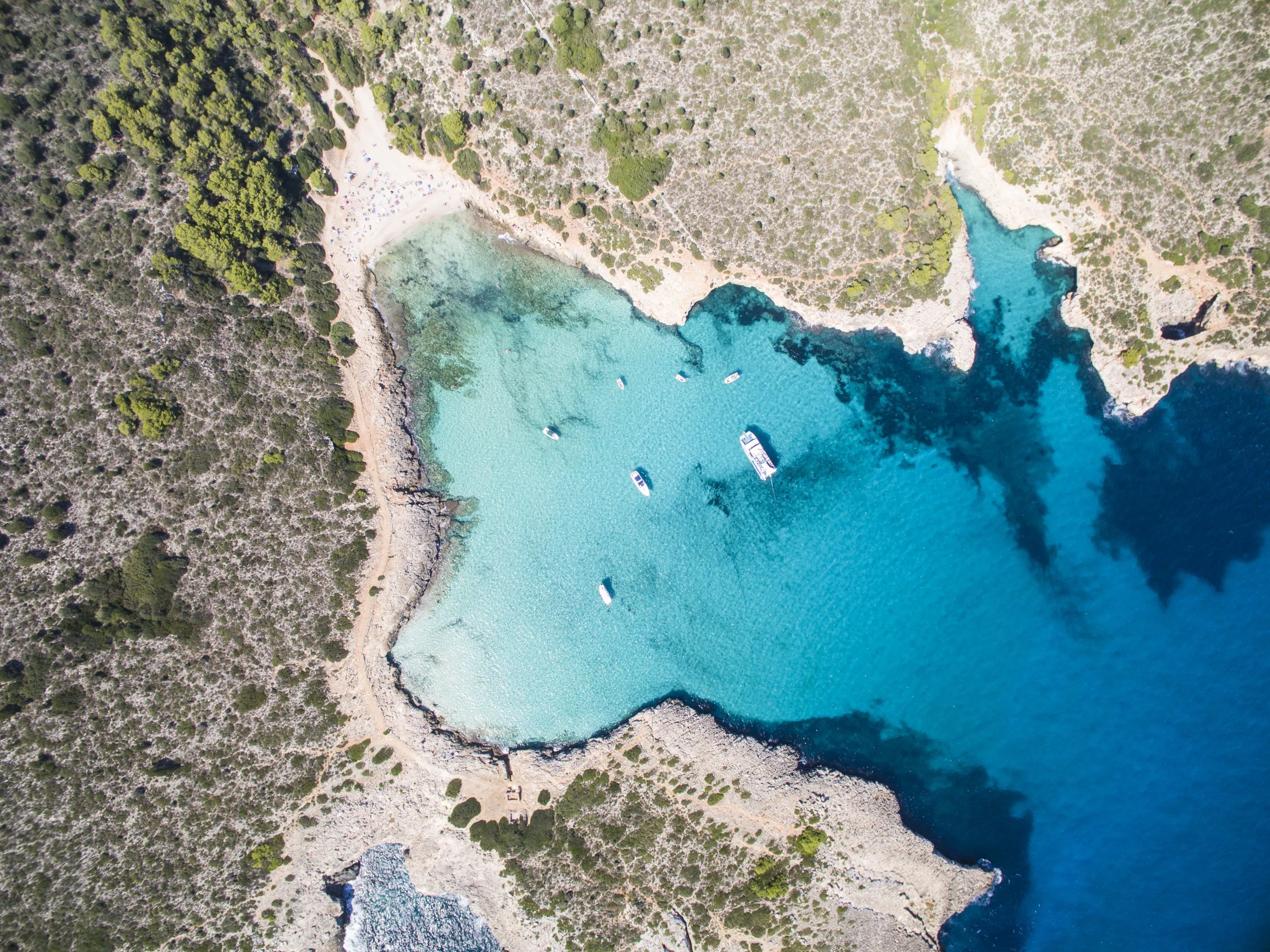 Aerial view of a small coastal bay of Cala Romantica Mallorca with turquoise water, surrounded by rocky and sandy shores, with boats anchored in the water and a wooded area nearby.