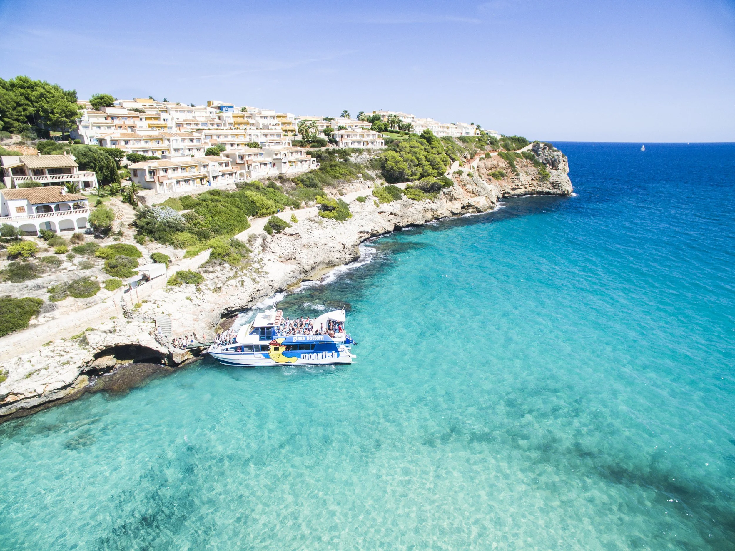 A boat named 'moonfish' with a glass bottom docked near rocky shore in turquoise water, with a hillside of white buildings and green trees in the background on a clear, sunny day at cala Romantica where the boat picks up passengers