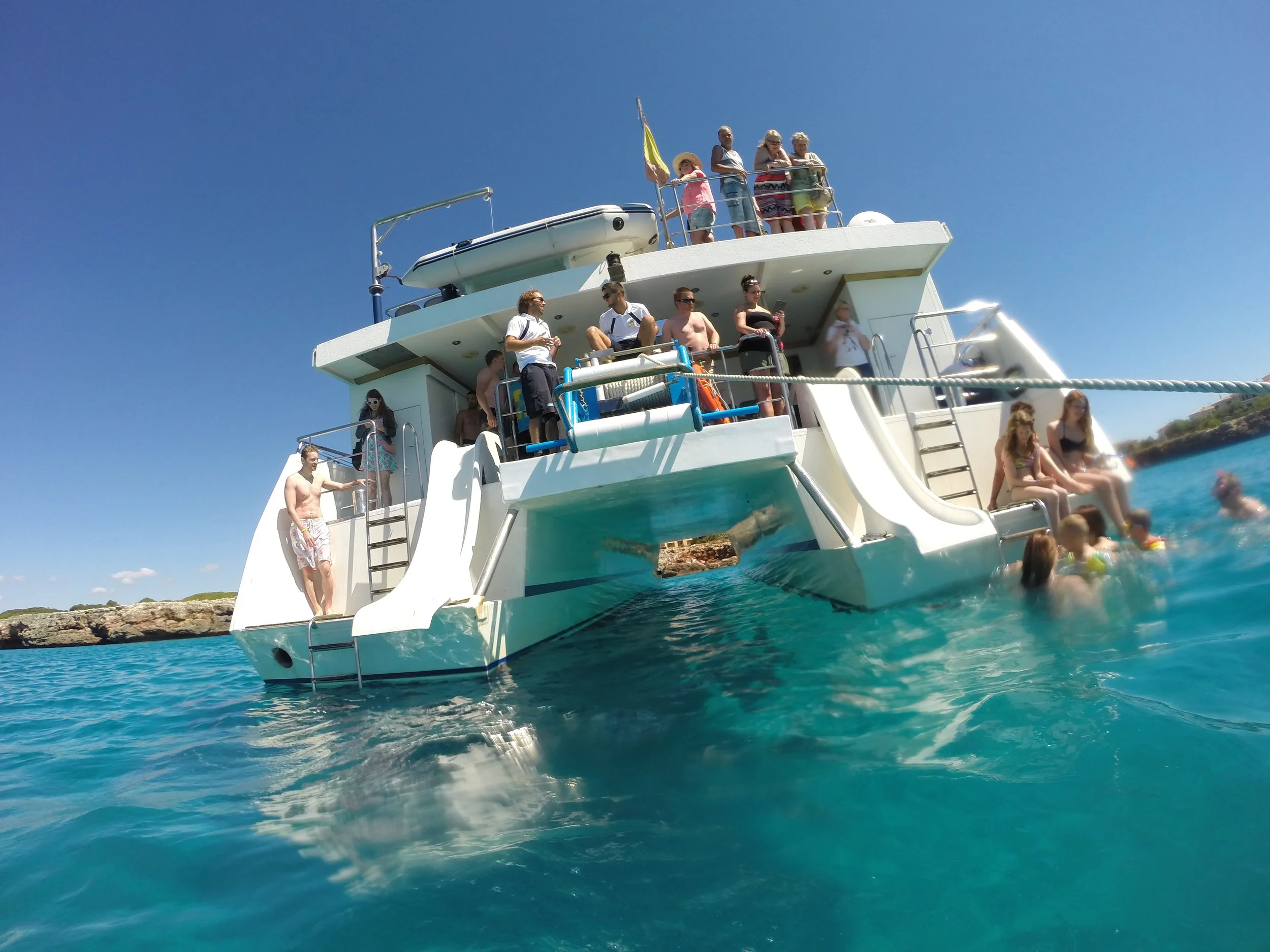People  swimming at Cala varques from Moonfish's glassbottom boat, some swimming in turquoise water, under a clear blue sky.